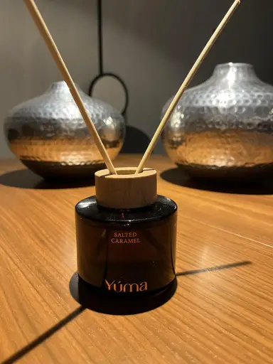 Reed diffuser Salted Caramel της YUMA με ξύλινο πώμα και δύο στικ σε ξύλινη επιφάνεια.