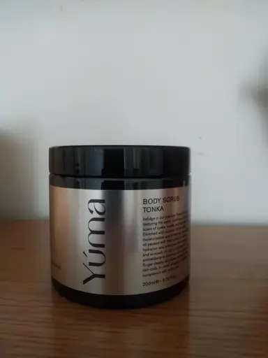 Ένα μαύρο βαζάκι Body Scrub Tonka της YUMA με ασημί ετικέτα πάνω σε ξύλινη επιφάνεια.