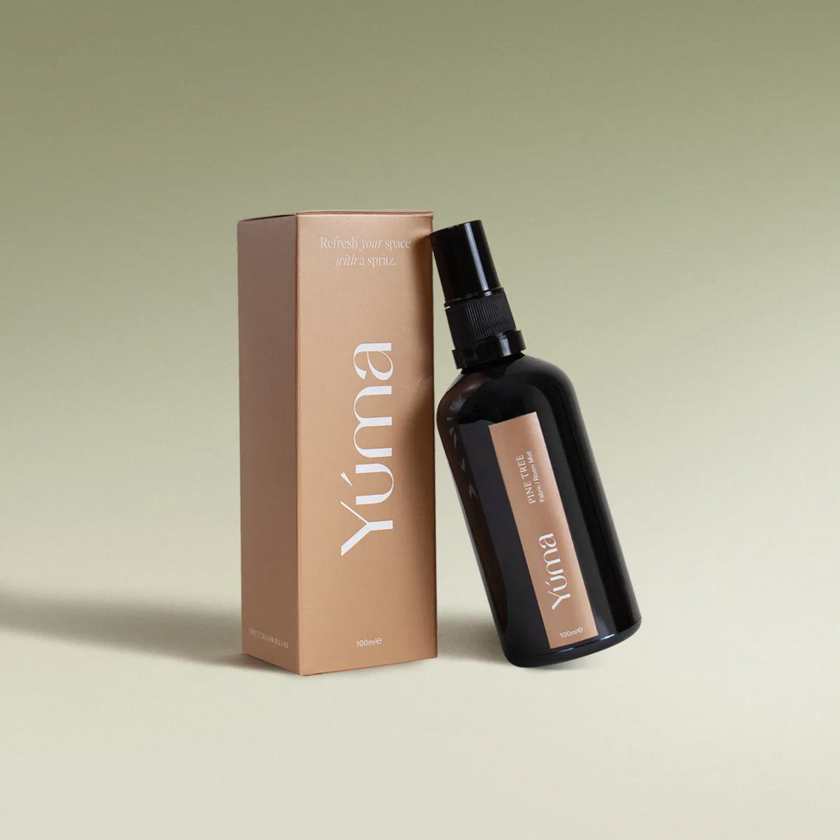 Σκούρο γυάλινο μπουκάλι Pine Tree Fabric Mist της YUMA δίπλα στην μπεζ χάρτινη συσκευασία του.