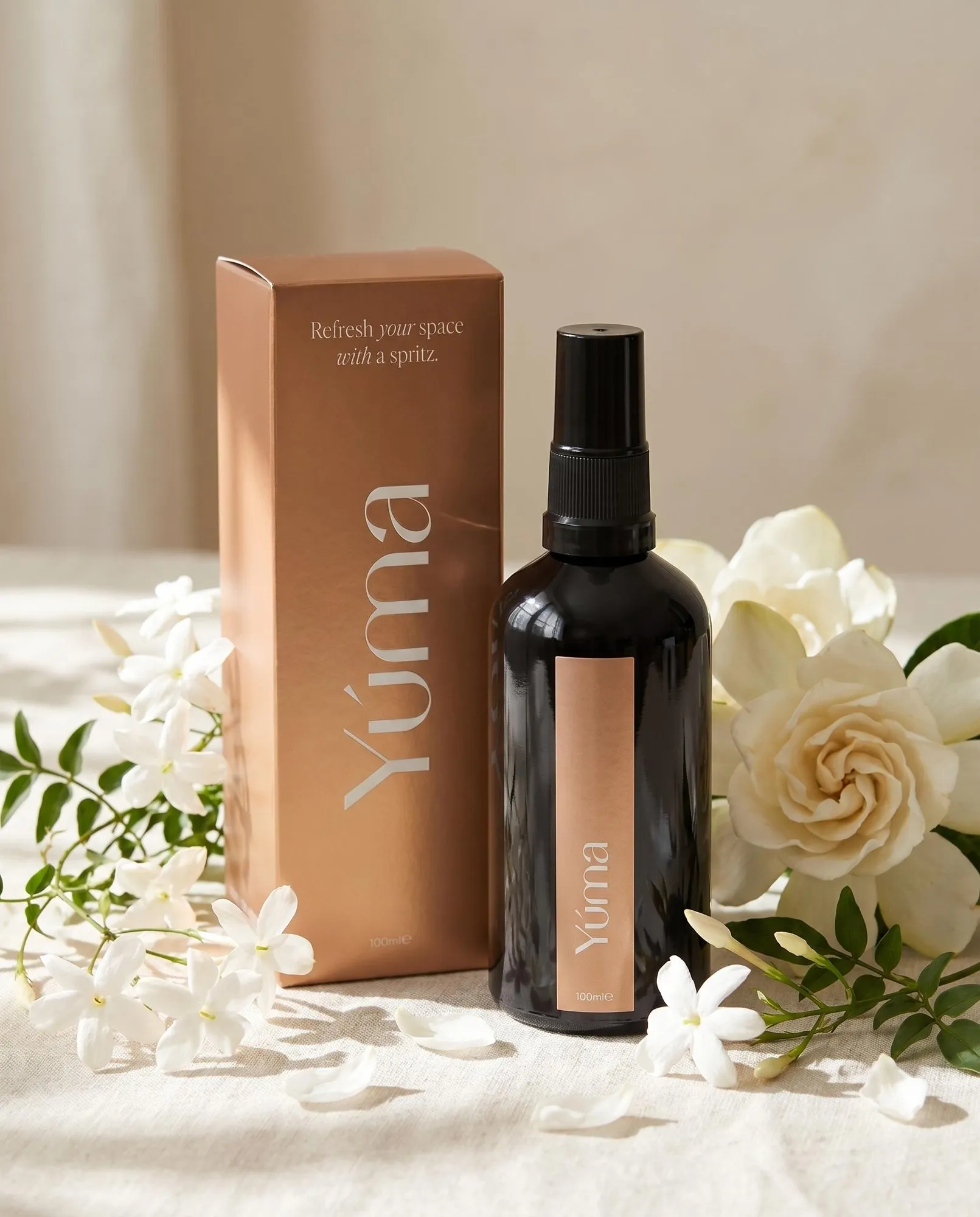 Μαύρο μπουκάλι fabric spray Eternal Eden της YUMA δίπλα σε χάλκινο κουτί και λευκά άνθη γιασεμιού.