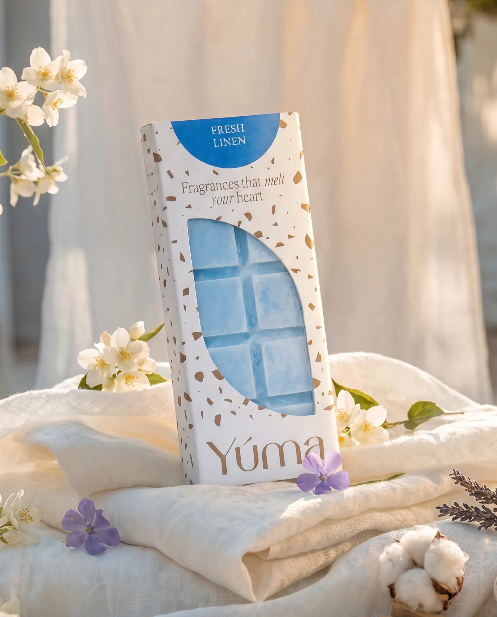 Μπλε Wax Melts Fresh Linen της YUMA σε λευκή συσκευασία πάνω σε λευκά υφάσματα με άνθη.