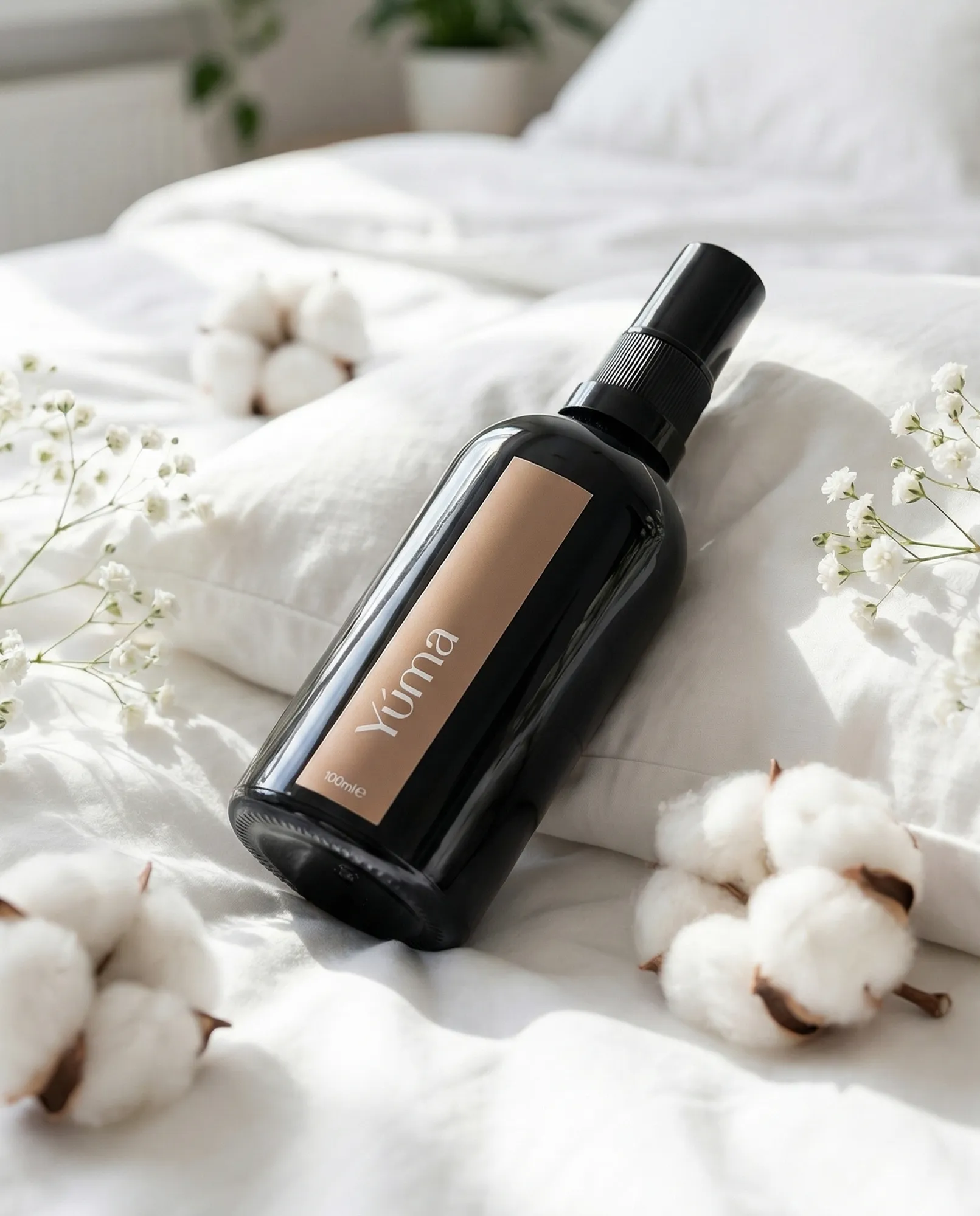 Σκούρο μπουκάλι Fresh Linen Fabric/Room Mist της YUMA πάνω σε λευκά κλινοσκεπάσματα, με άνθη βαμβακιού και γυψοφύλλη.