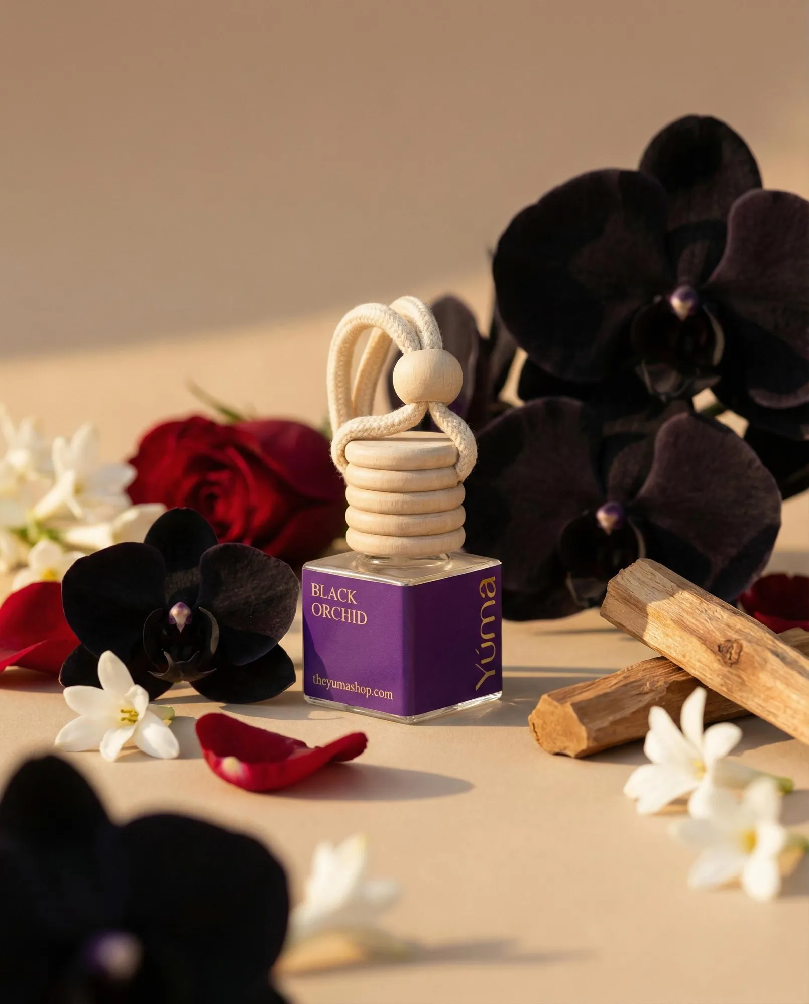 Car Diffuser της YUMA με άρωμα Black Orchid ανάμεσα σε μαύρες ορχιδέες, κόκκινα τριαντάφυλλα και ξύλα.