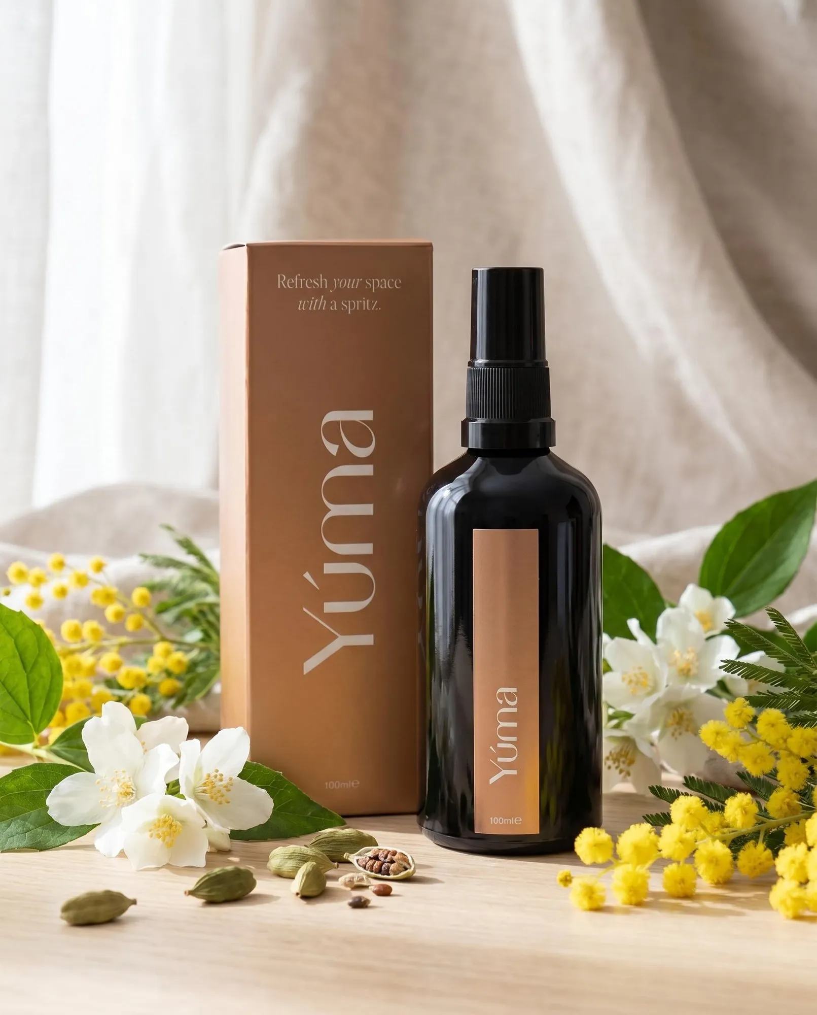 Μαύρο μπουκάλι fabric spray Oriental Mimosa της YUMA δίπλα σε χάρτινη συσκευασία, λευκά άνθη και μιμόζες.
