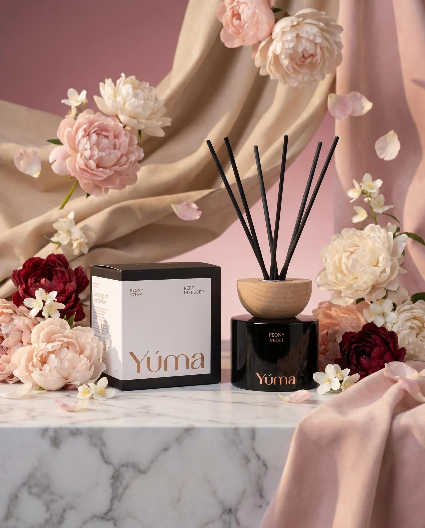 Reed diffuser Peony Velvet της YUMA σε μαύρο βάζο δίπλα στη συσκευασία του με φόντο παιώνιες.