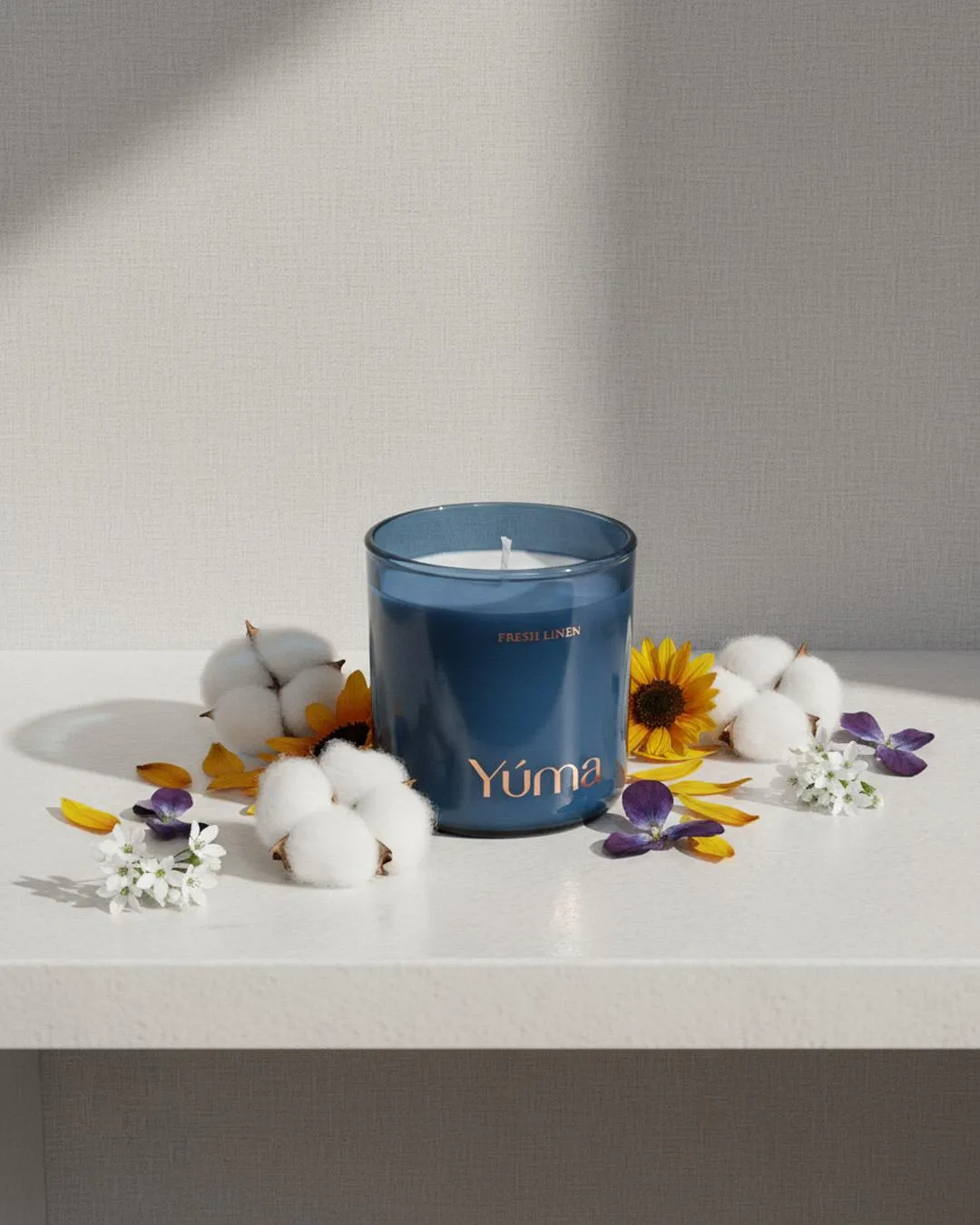 Μπλε γυάλινο κερί Fresh Linen της YUMA ανάμεσα σε άνθη βαμβακιού και κίτρινα πέταλα.