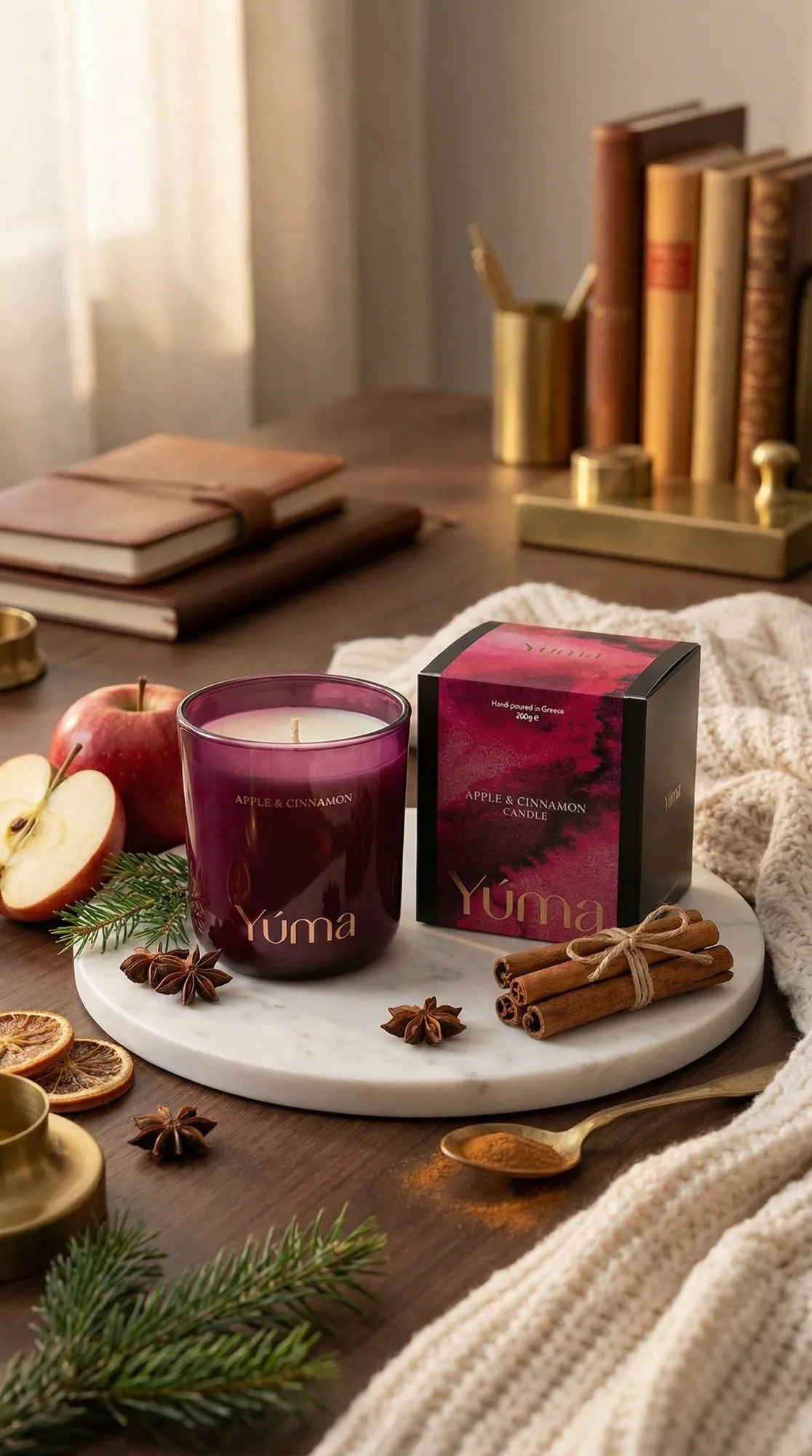 Κερί Apple Cinnamon της YUMA σε καπνιστό ροζ γυάλινο δοχείο δίπλα σε μήλα και ξυλάκια κανέλας.
