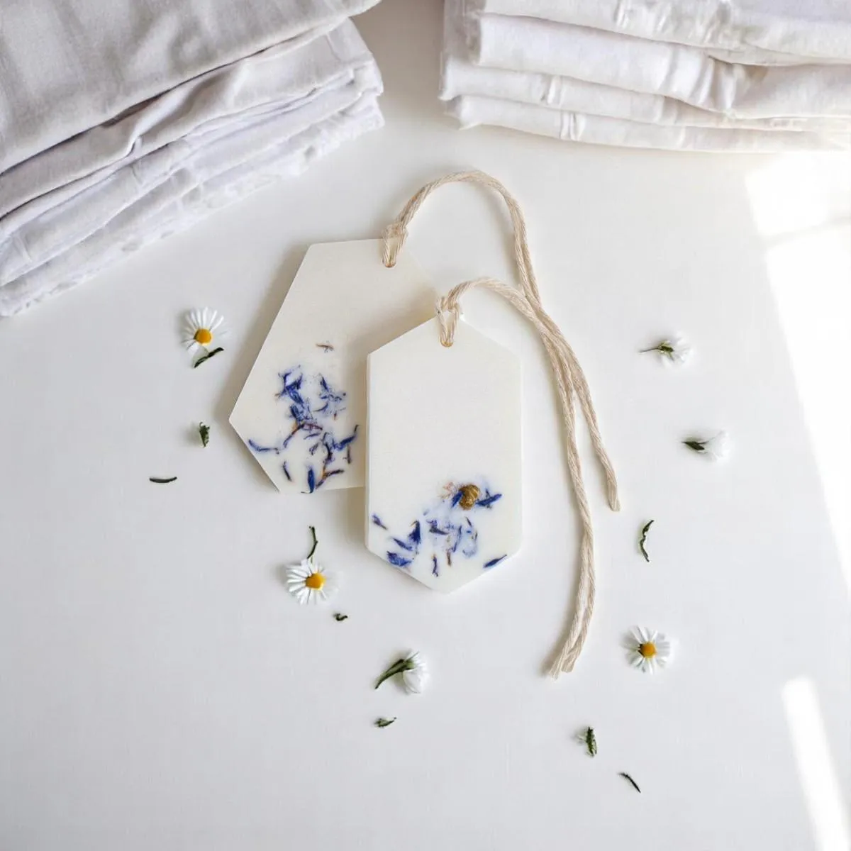 Δύο λευκά Fresh Linen Wax Tablets με μπλε άνθη δίπλα σε διπλωμένα υφάσματα.