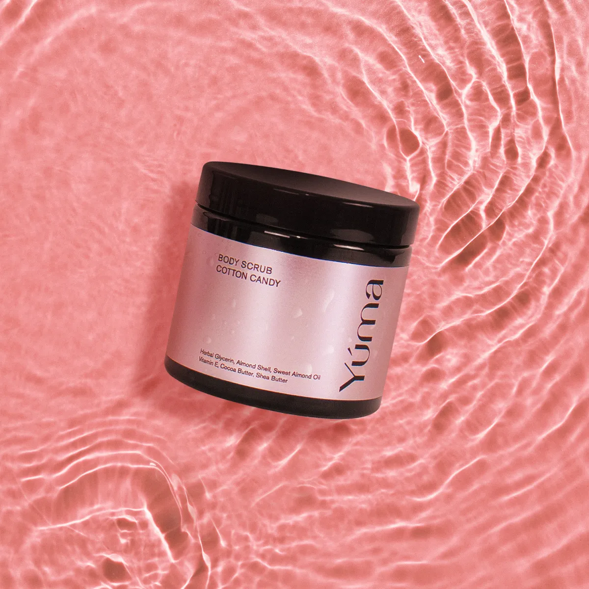 Body Scrub Cotton Candy της YUMA σε σκούρο βάζο πάνω σε ροζ νερό με κυματισμούς.