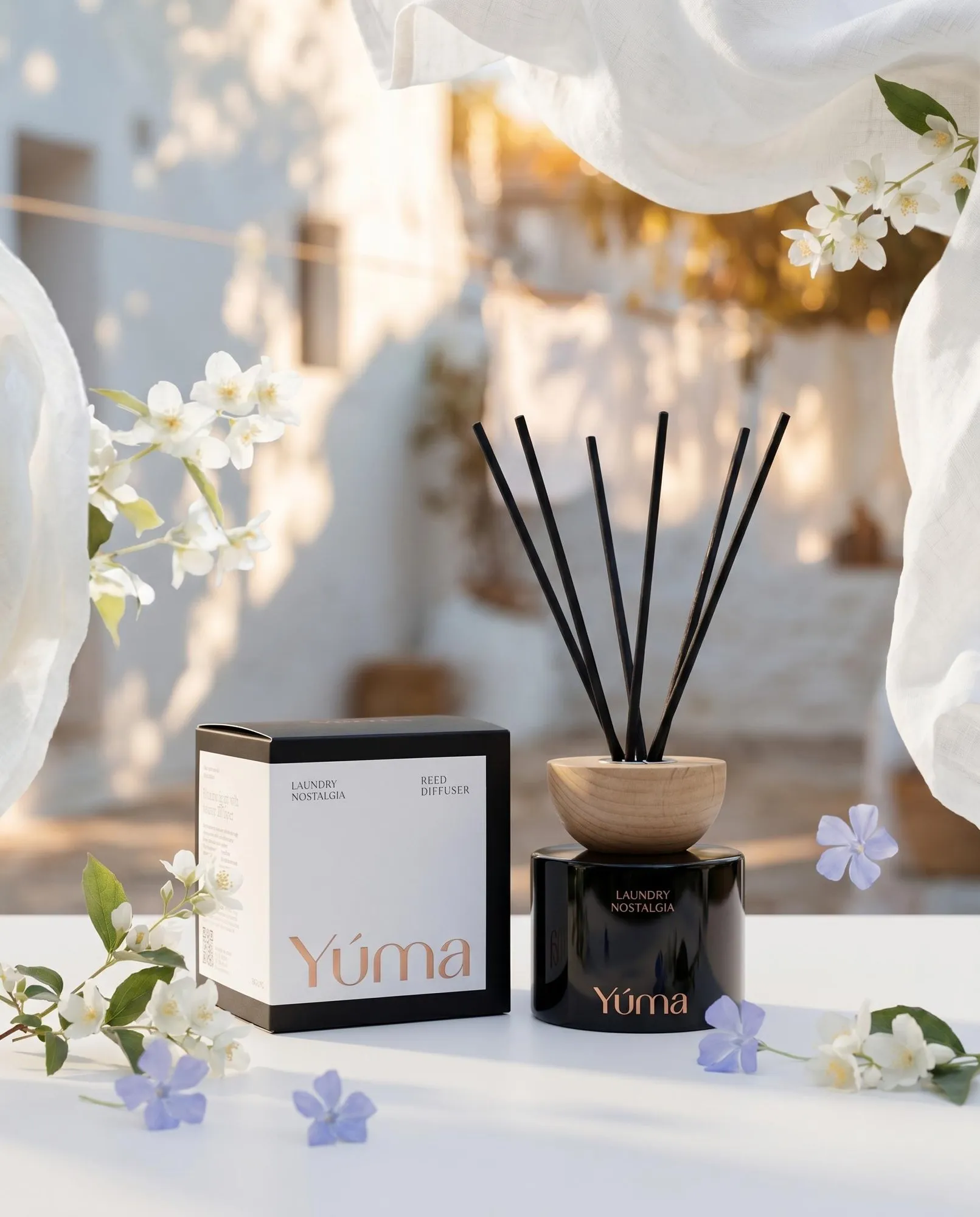 Reed diffuser Laundry Nostalgia της YUMA δίπλα στη συσκευασία του, ανάμεσα σε λευκά και μοβ άνθη.