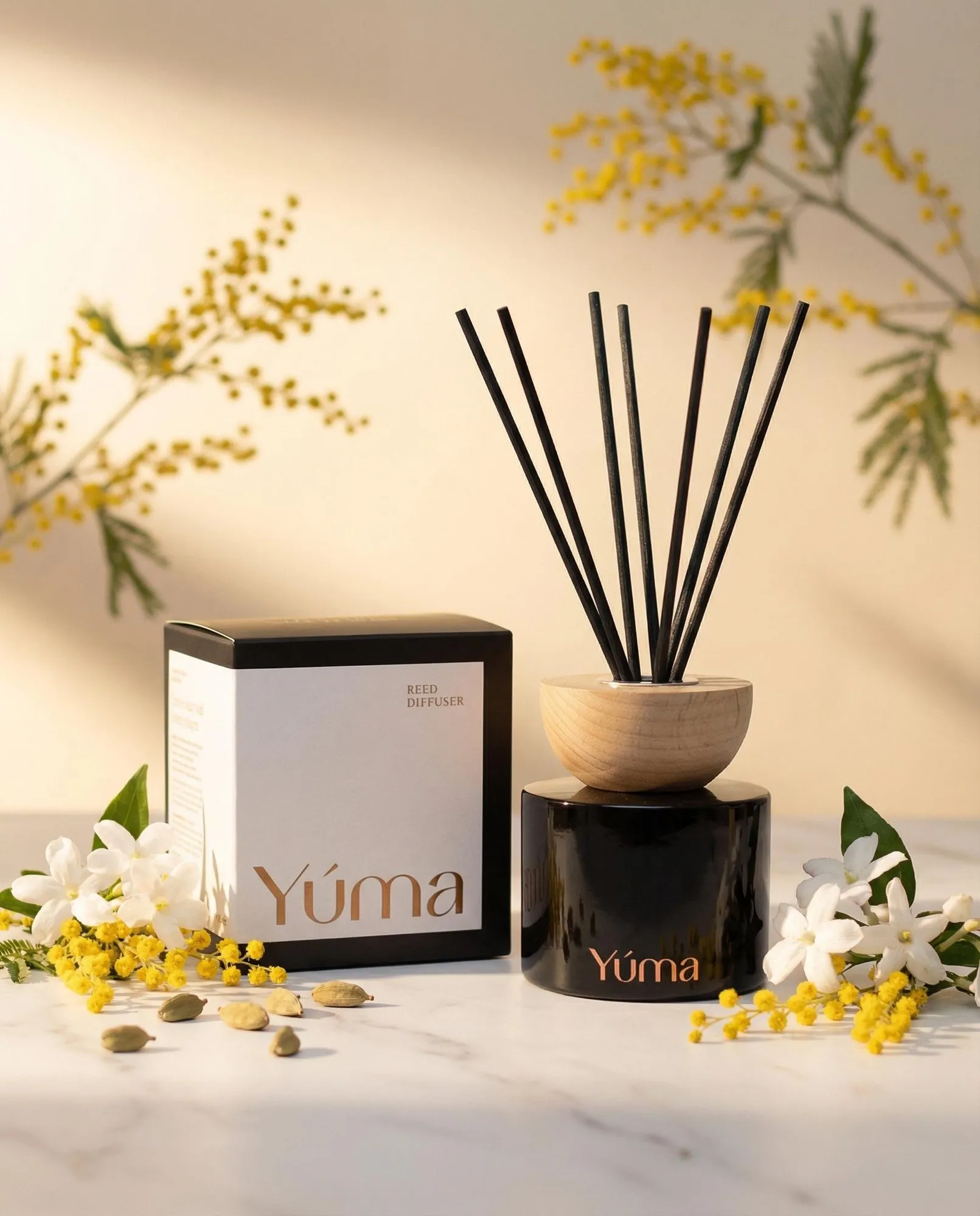 Reed diffuser της YUMA με άρωμα Oriental Mimosa δίπλα σε λευκά άνθη και κίτρινη μιμόζα.