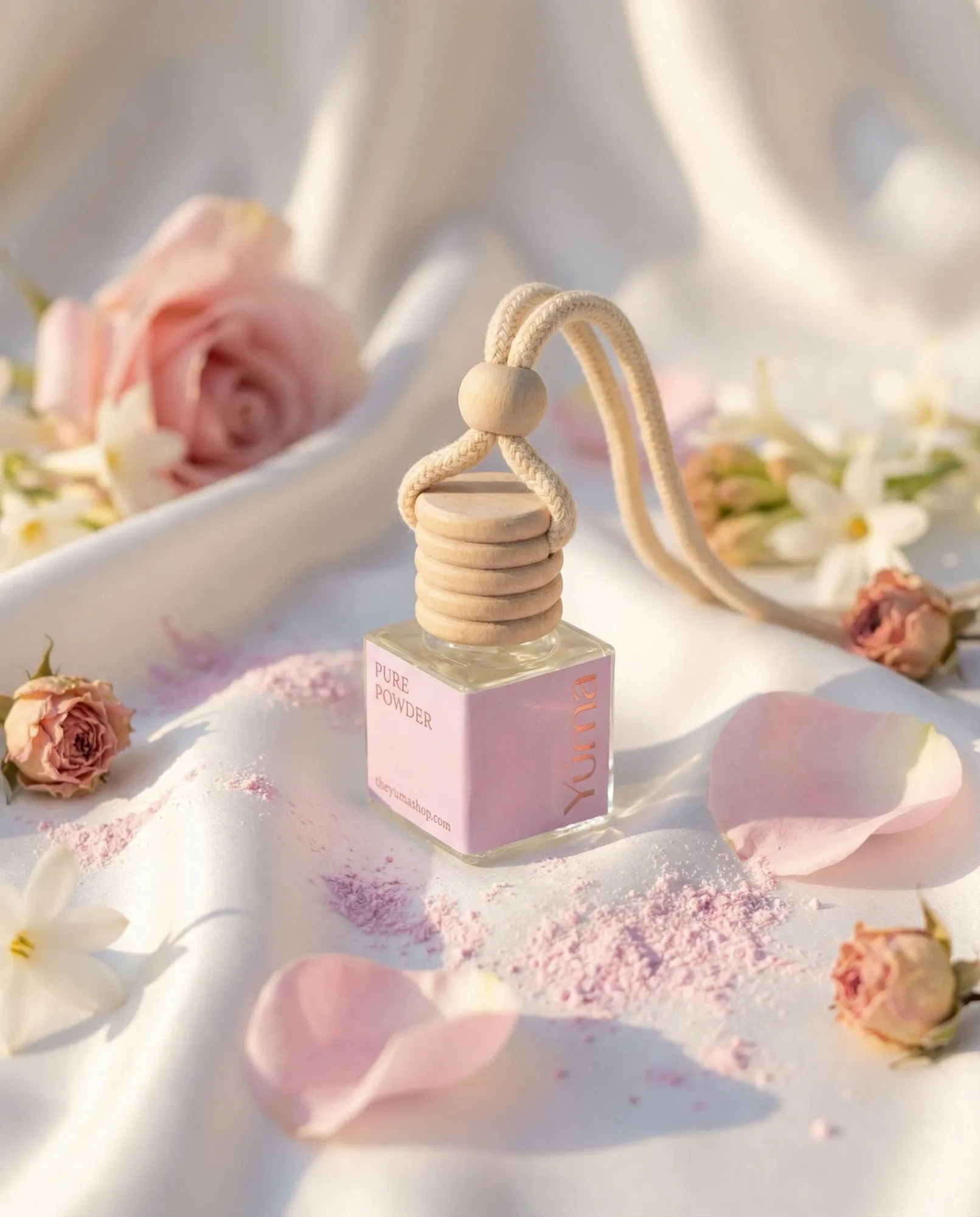 Car Diffuser Pure Powder της YUMA πάνω σε λευκό ύφασμα με ροζ πέταλα και άνθη.