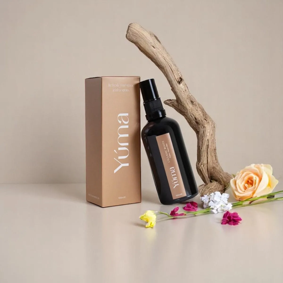 Σκούρο μπουκάλι Fabric Mist Cycladic Roots της YUMA δίπλα σε χάρτινη συσκευασία, ξύλο και λουλούδια.