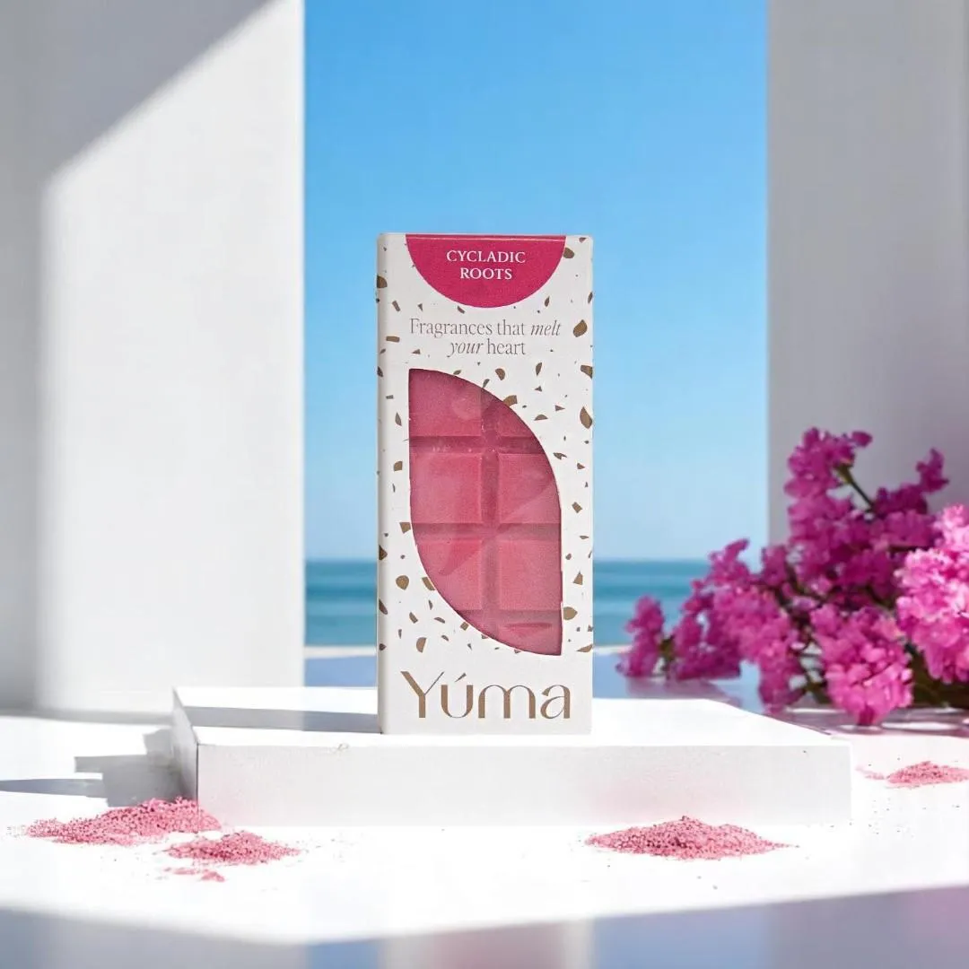 Ροζ Cycladic Roots wax melts της YUMA σε λευκή συσκευασία με φόντο τη θάλασσα και λουλούδια.