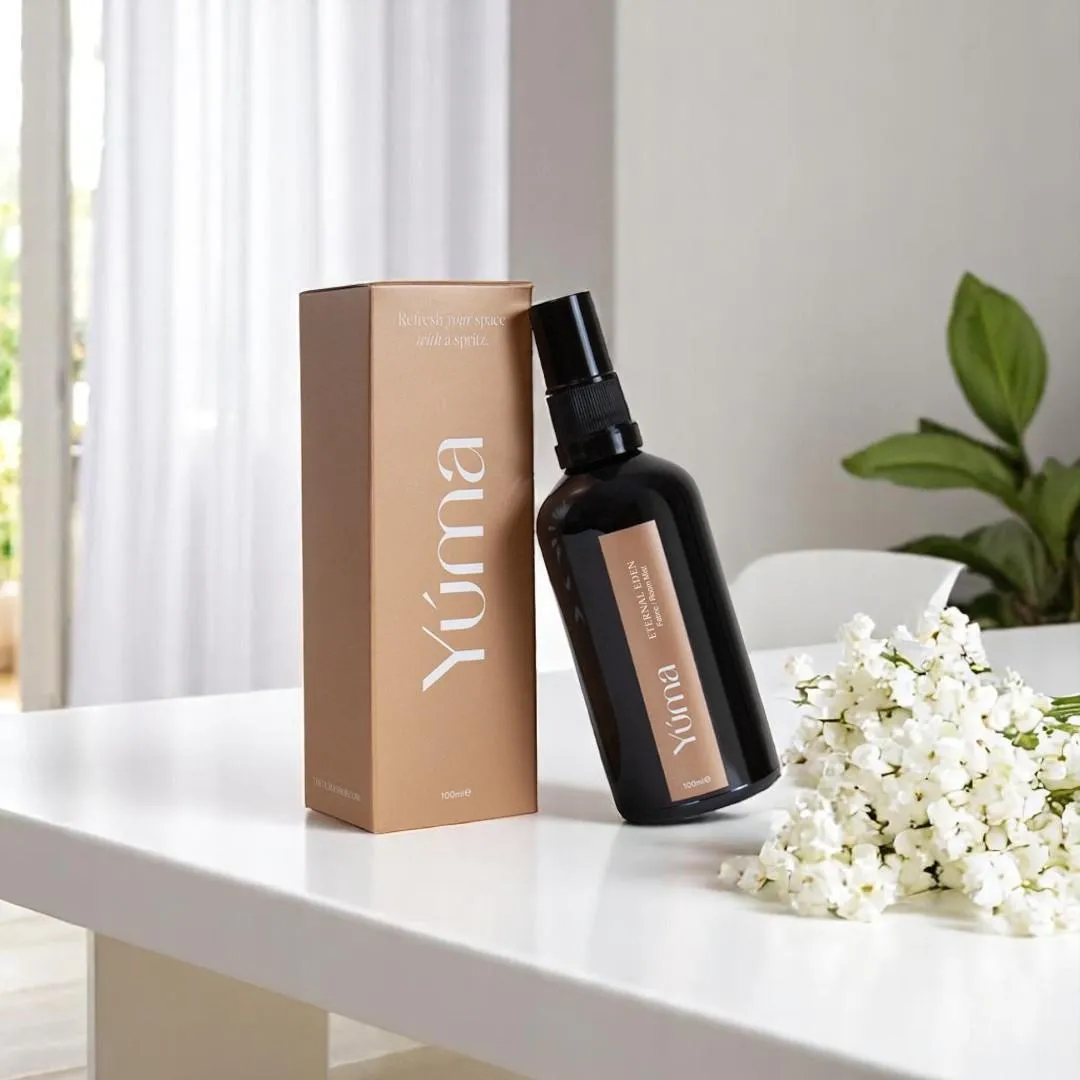 Room spray Eternal Eden της YUMA σε σκούρο μπουκάλι δίπλα στη συσκευασία του και λευκά άνθη.