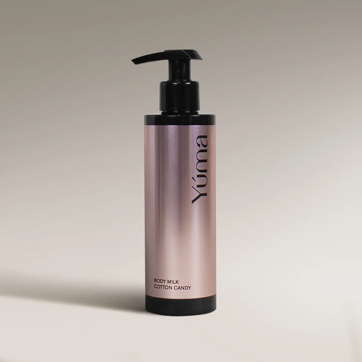 Μεταλλικό ροζ μπουκάλι Body Milk Cotton Candy της YUMA με μαύρη αντλία σε μπεζ φόντο.