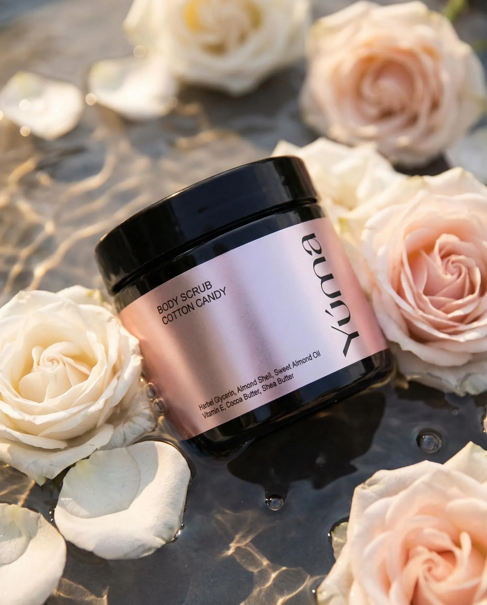 Body Scrub Cotton Candy της YUMA σε ροζ συσκευασία, μέσα σε νερό με ανοιχτόχρωμα τριαντάφυλλα.