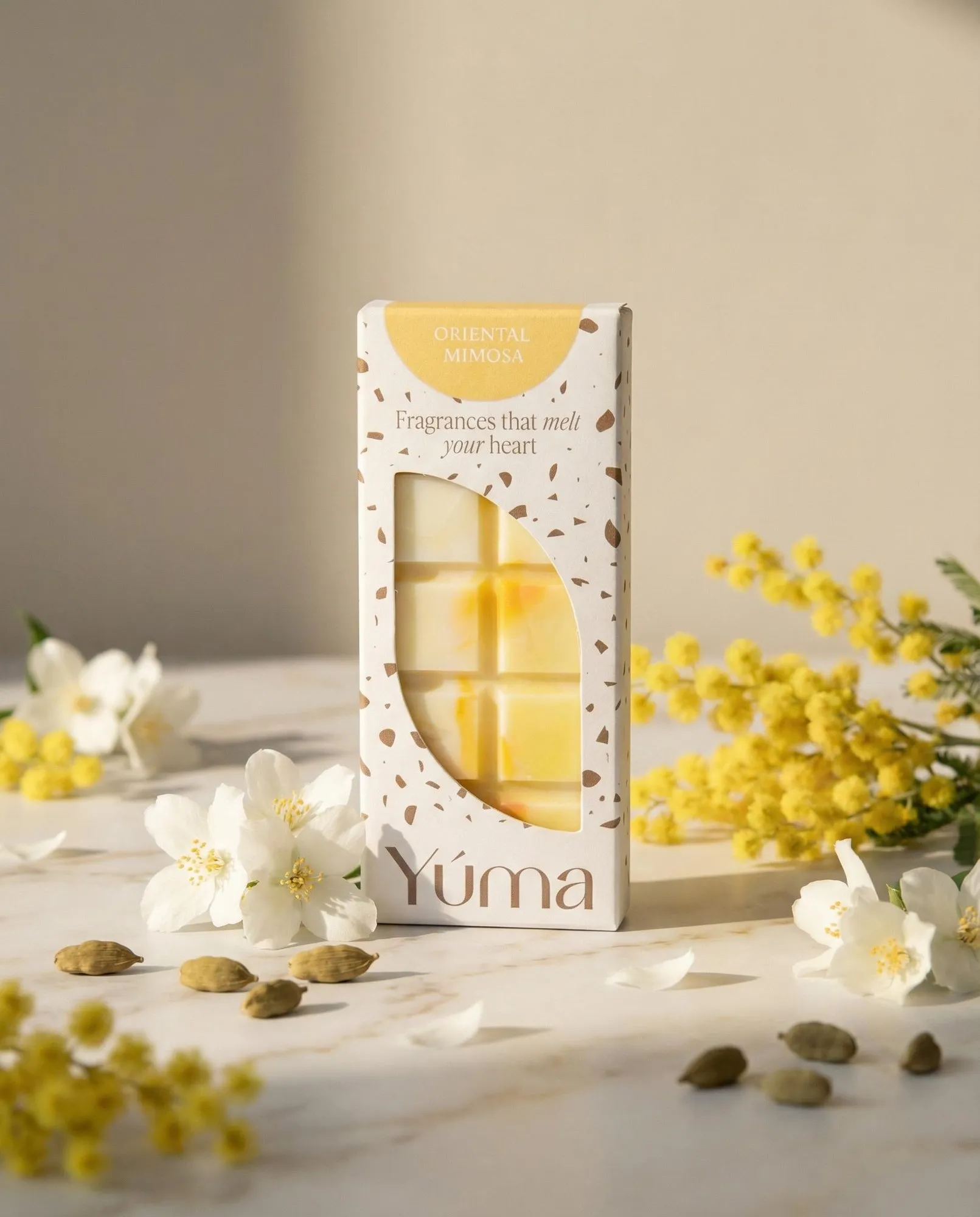 Συσκευασία Oriental Mimosa wax melts της YUMA ανάμεσα σε κίτρινα άνθη μιμόζας και λευκά λουλούδια.