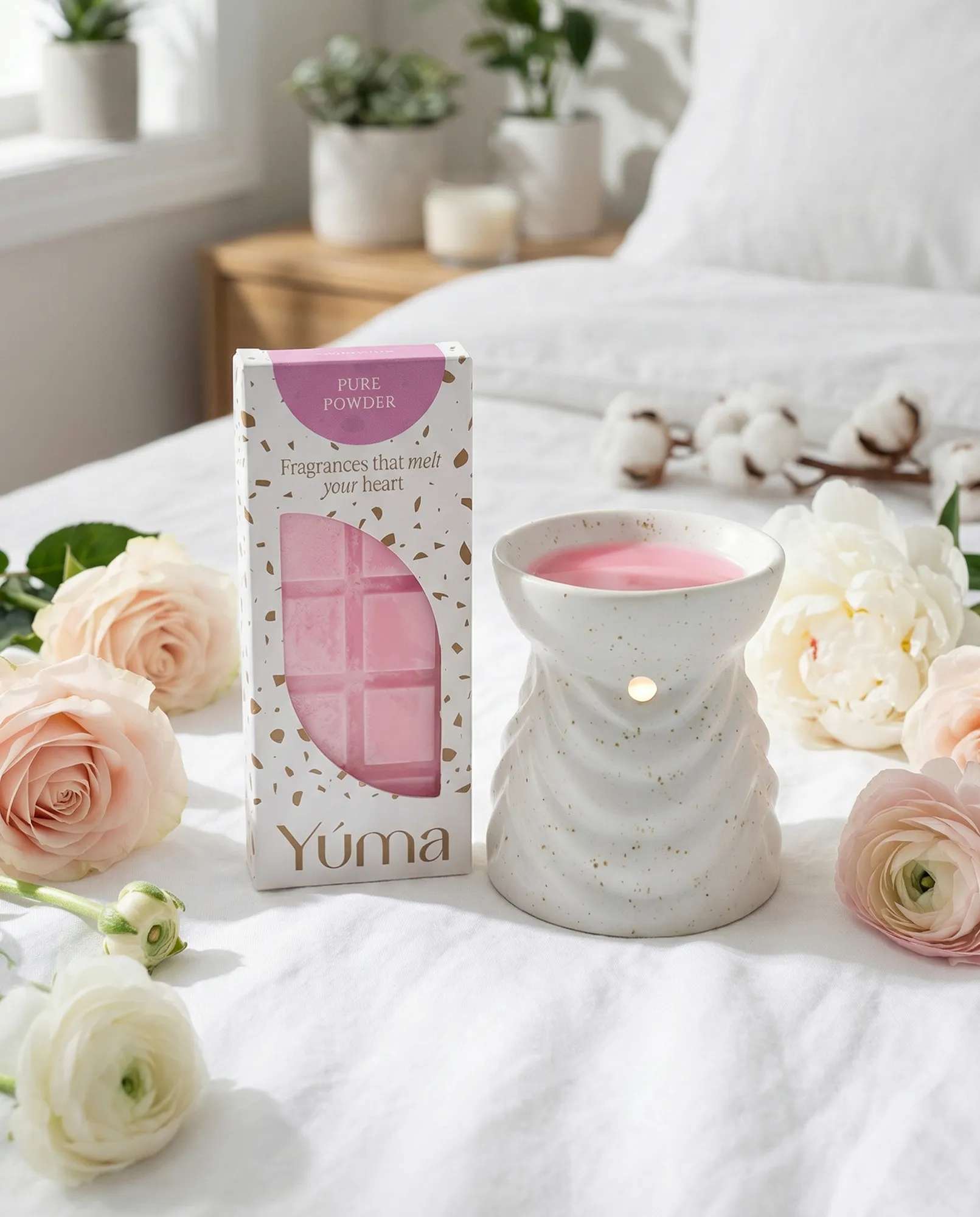 Ροζ Pure Powder wax melts της YUMA δίπλα σε κεραμικό αρωματιστή και τριαντάφυλλα πάνω σε λευκό κρεβάτι.