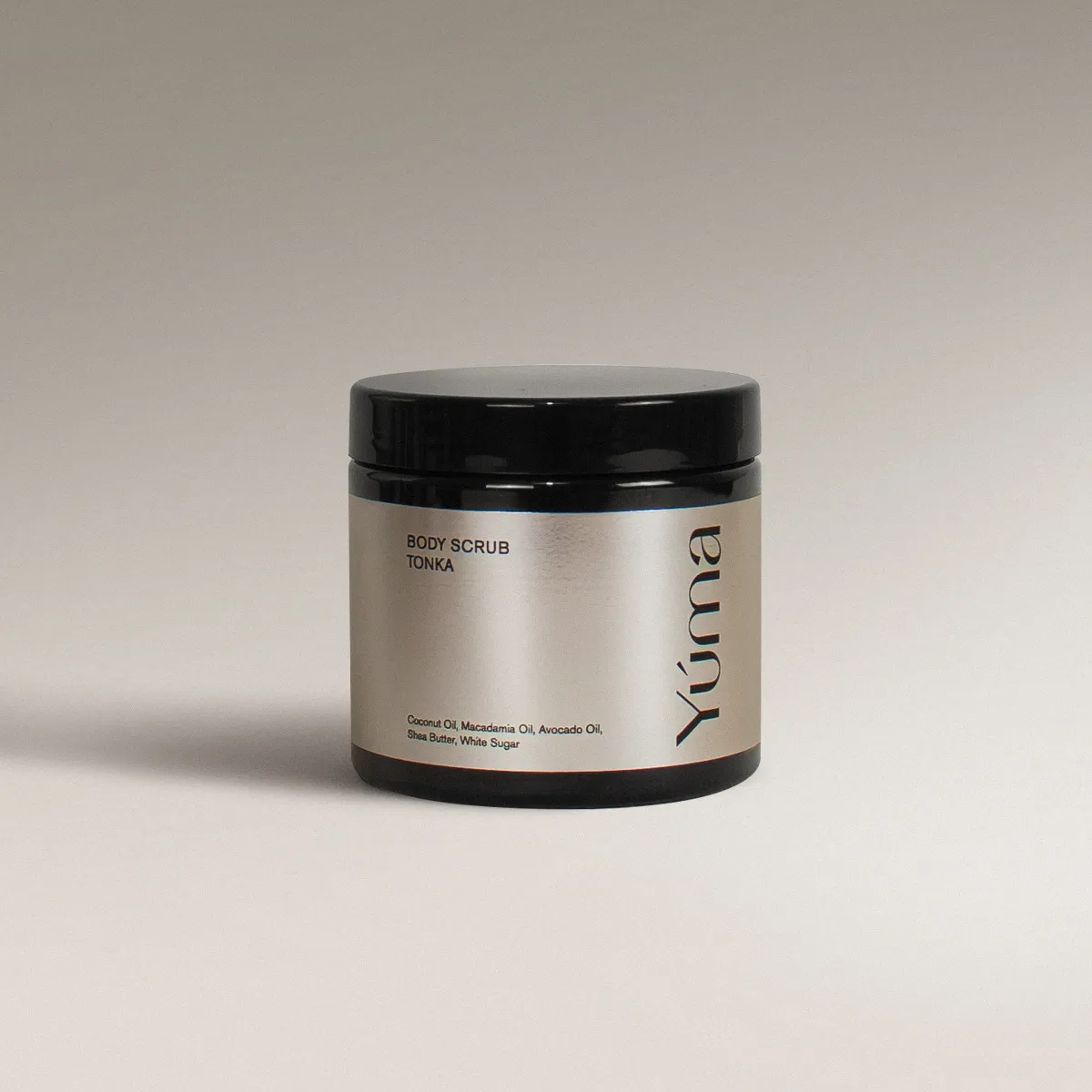 Μαύρο βάζο Body Scrub της YUMA με άρωμα Tonka και ασημί ετικέτα σε γκρίζο φόντο.