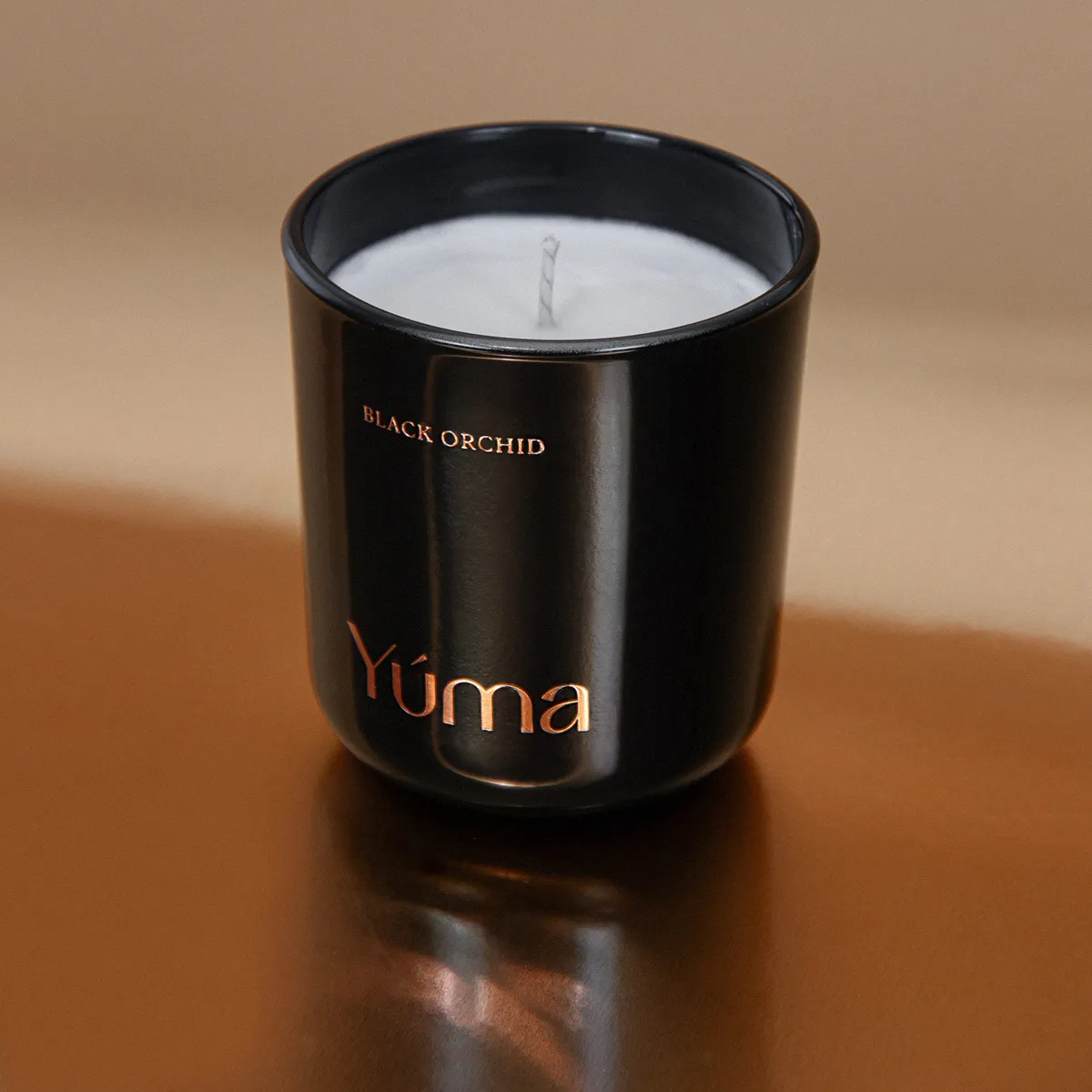 Μαύρο γυάλινο κερί Black Orchid της YUMA δίπλα σε λευκό κερί πάνω σε μπρούτζινη επιφάνεια.