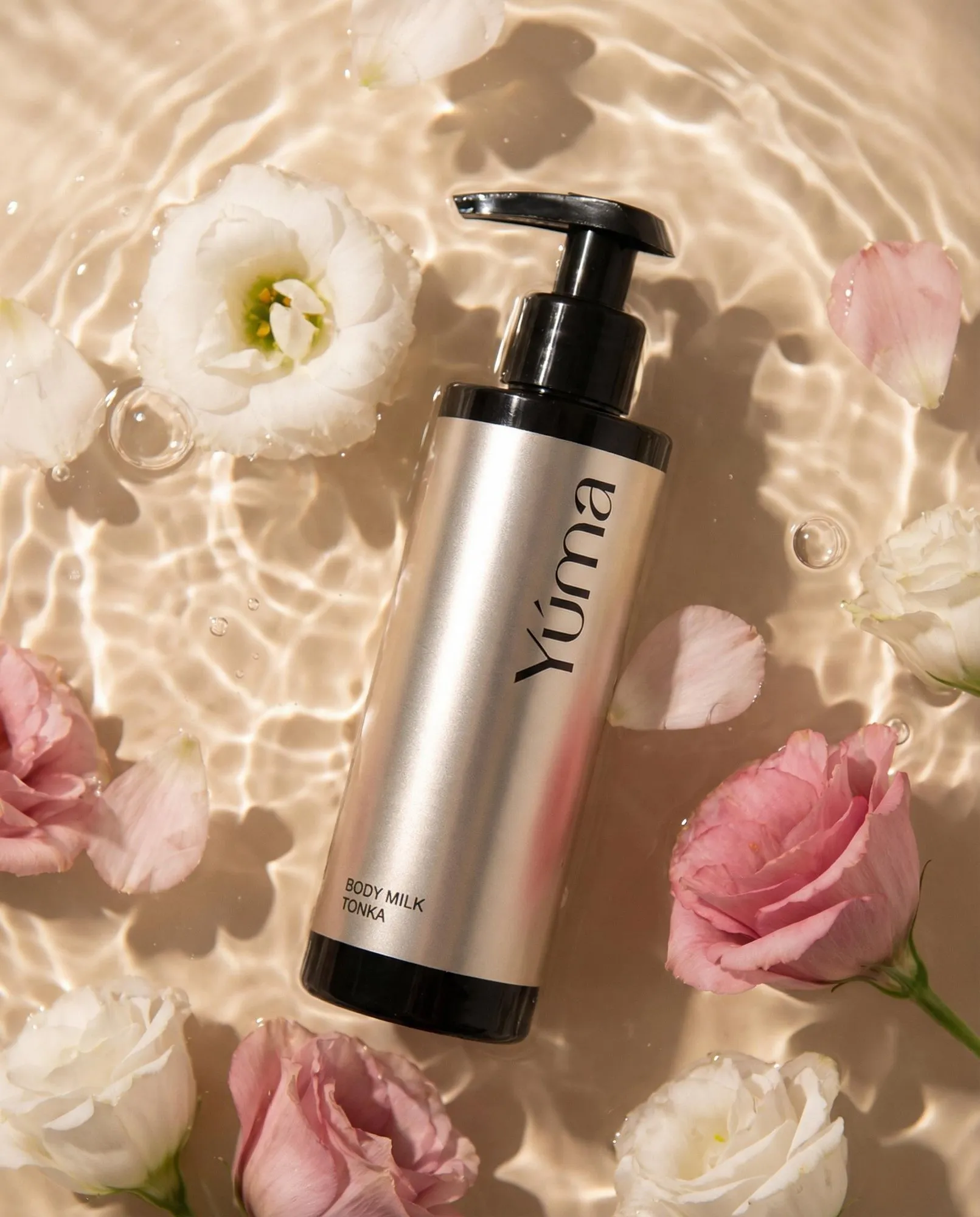 Body Milk Tonka της YUMA σε μεταλλικό μπουκάλι μέσα σε νερό με ροζ και λευκά άνθη.