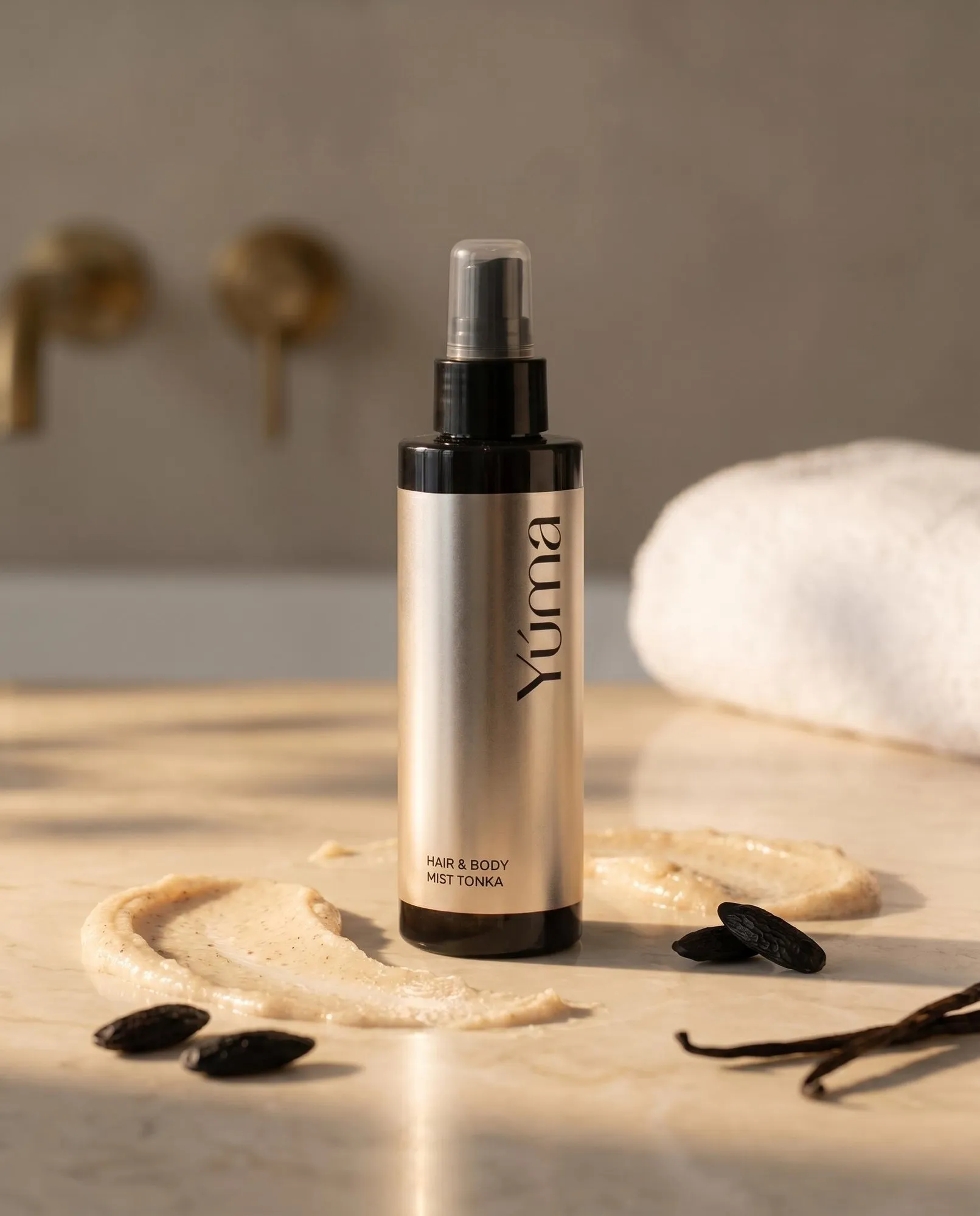 Hair & Body Mist Tonka της YUMA σε χρυσό μπουκάλι δίπλα σε κρέμα σώματος και σπόρους ως οπτικές αναφορές στο άρωμα.