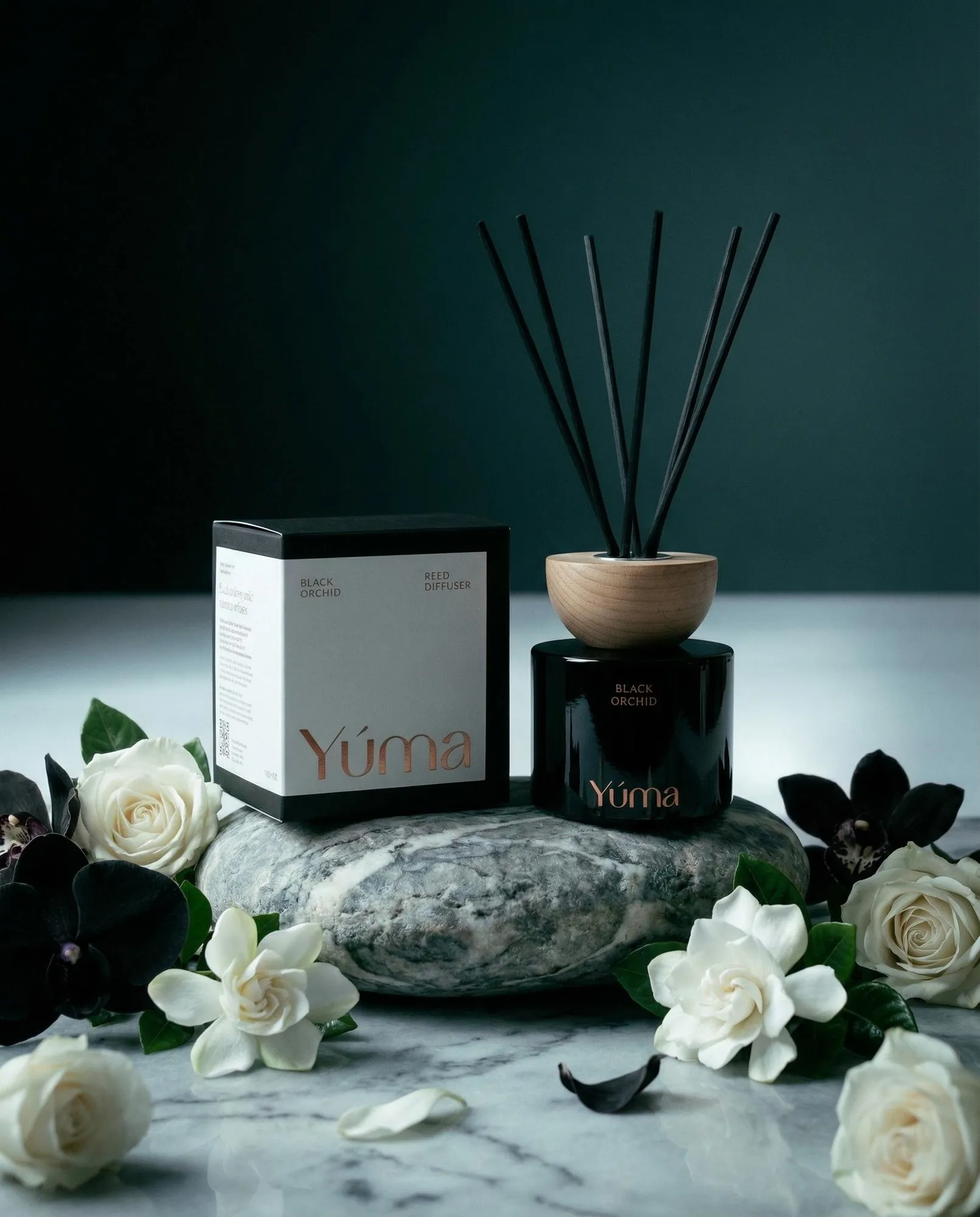 Το Black Orchid reed diffuser της YUMA δίπλα στη συσκευασία του πάνω σε πέτρα με λευκά άνθη.