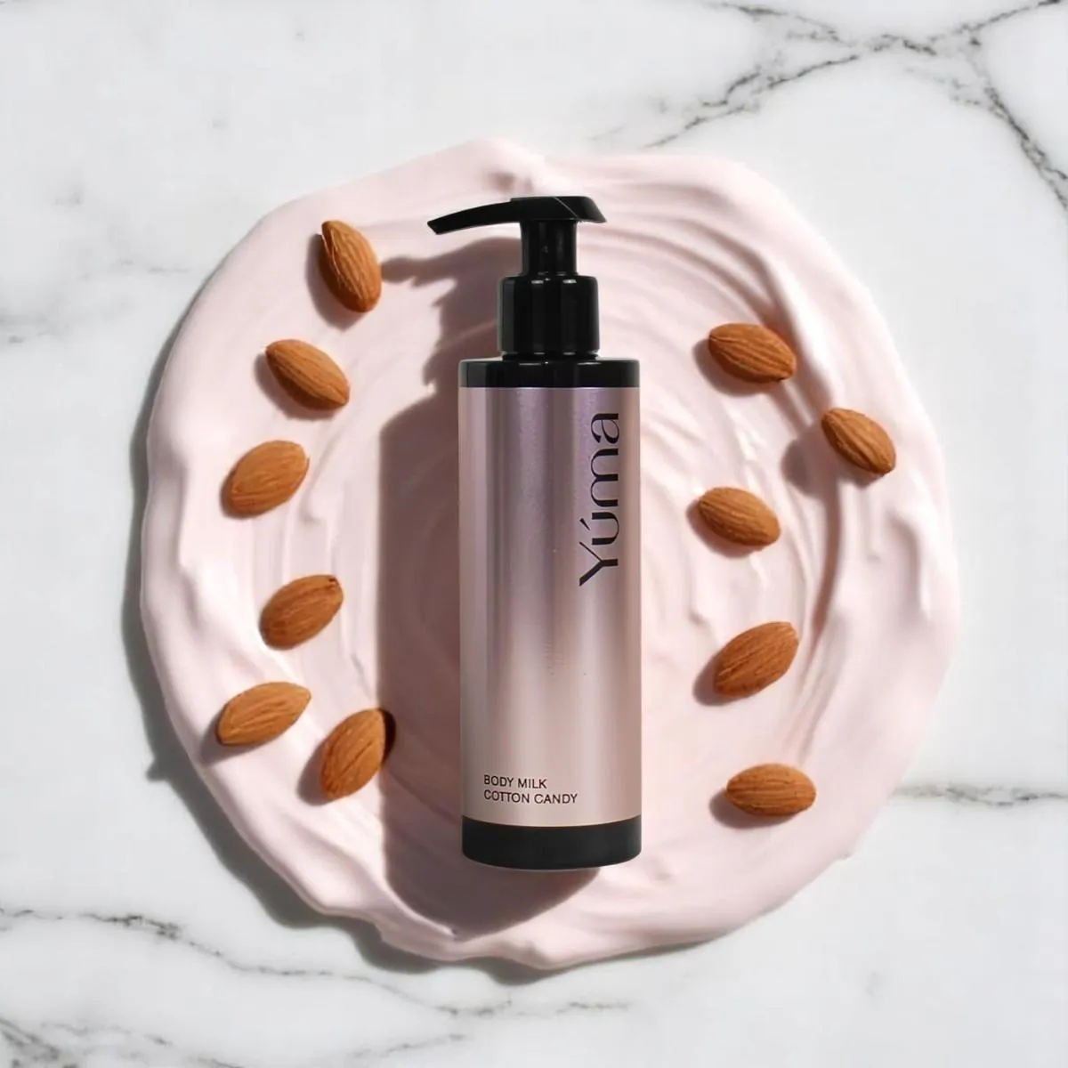 Body Milk Cotton Candy της YUMA πάνω σε ροζ κρέμα με αμύγδαλα σε μαρμάρινη επιφάνεια.