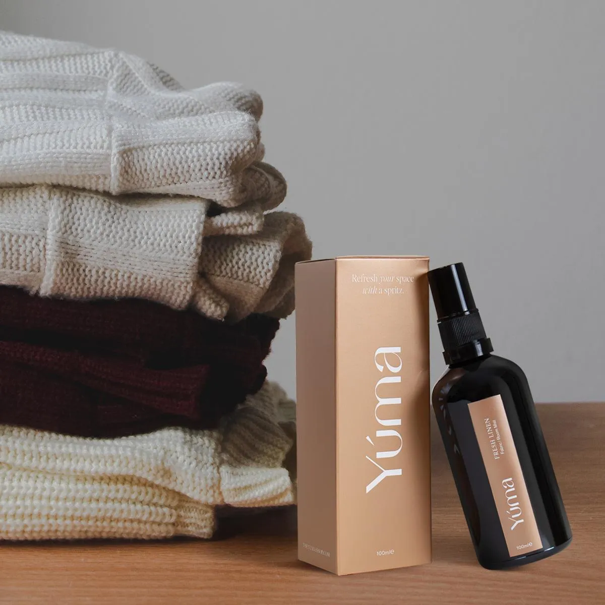 Σκούρο μπουκάλι Fabric Mist της YUMA δίπλα σε στοίβα από πλεκτά υφάσματα πάνω σε ξύλινη επιφάνεια.
