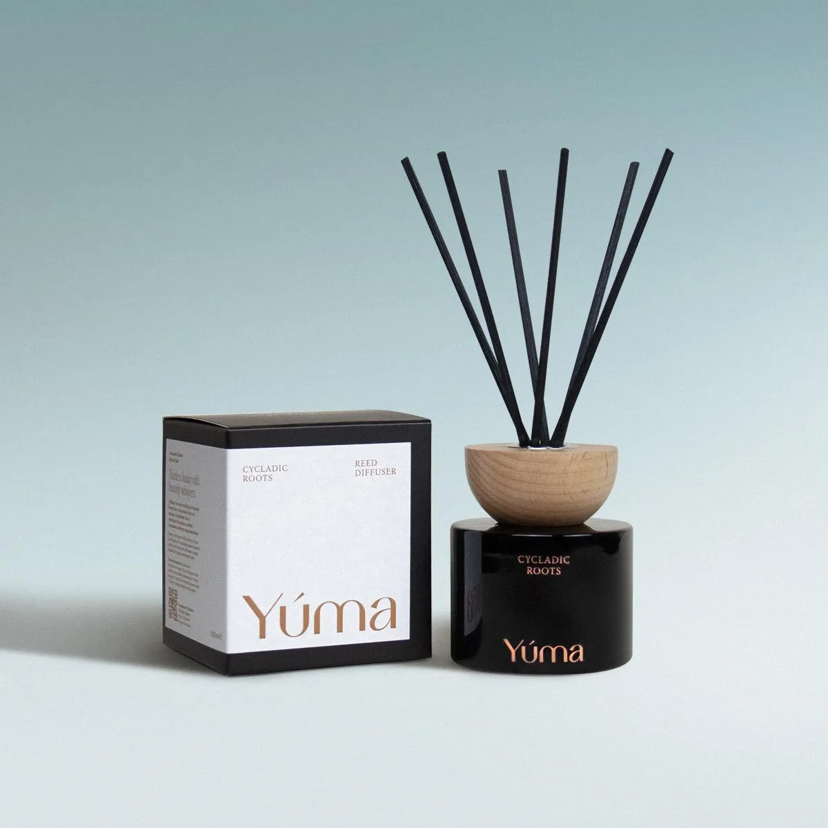 Μαύρο γυάλινο reed diffuser Cycladic Roots της YUMA με ξύλινο καπάκι δίπλα στη συσκευασία του.