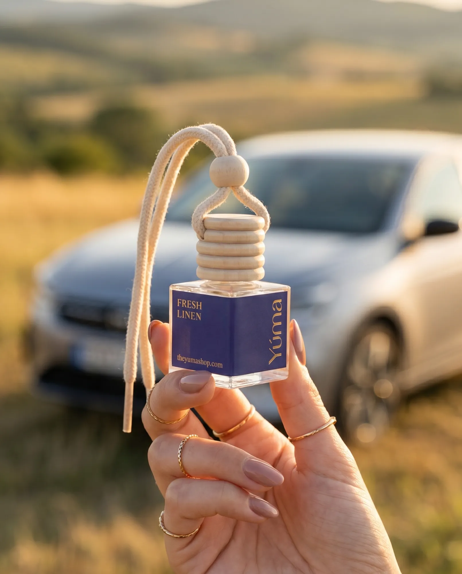 Γυναικείο χέρι κρατά το Car Diffuser Fresh Linen της YUMA με φόντο ένα αυτοκίνητο στη φύση.