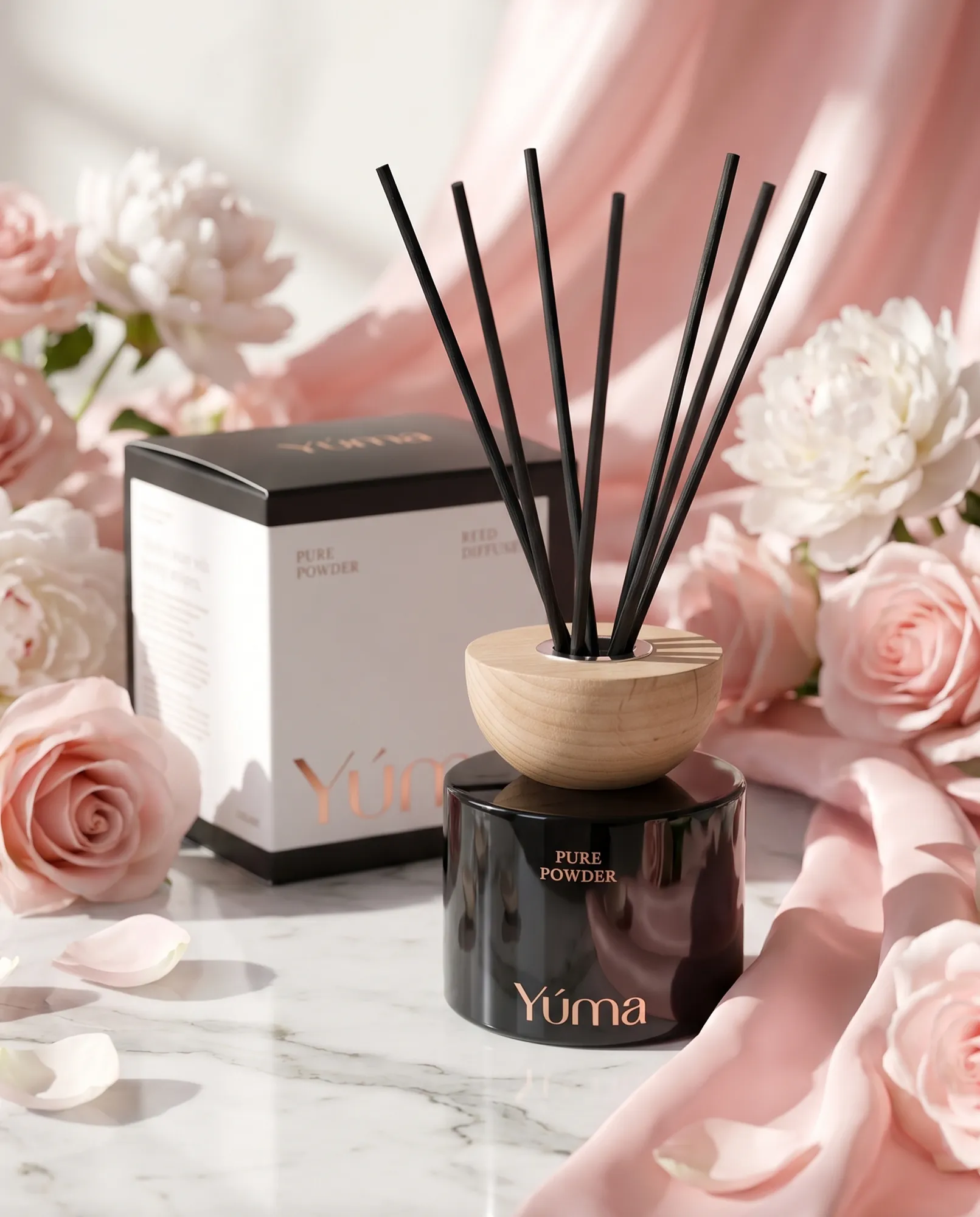 Reed diffuser Pure Powder της YUMA σε μαύρο γυάλινο δοχείο ανάμεσα σε ροζ τριαντάφυλλα και υφάσματα.