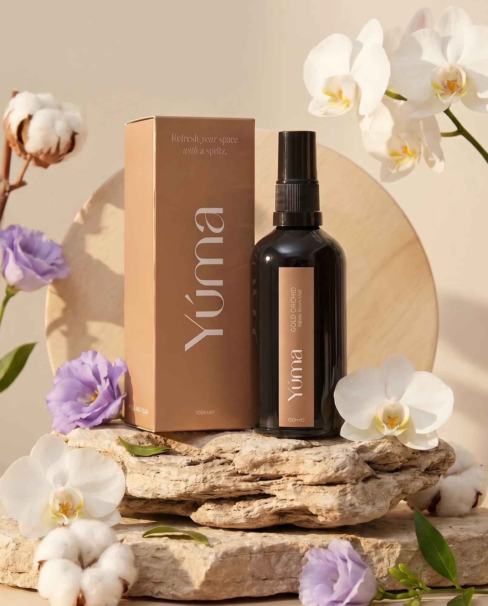 Το Fabric Room Mist Gold Orchid της YUMA δίπλα στη συσκευασία του, περιτριγυρισμένο από λευκές ορχιδέες.