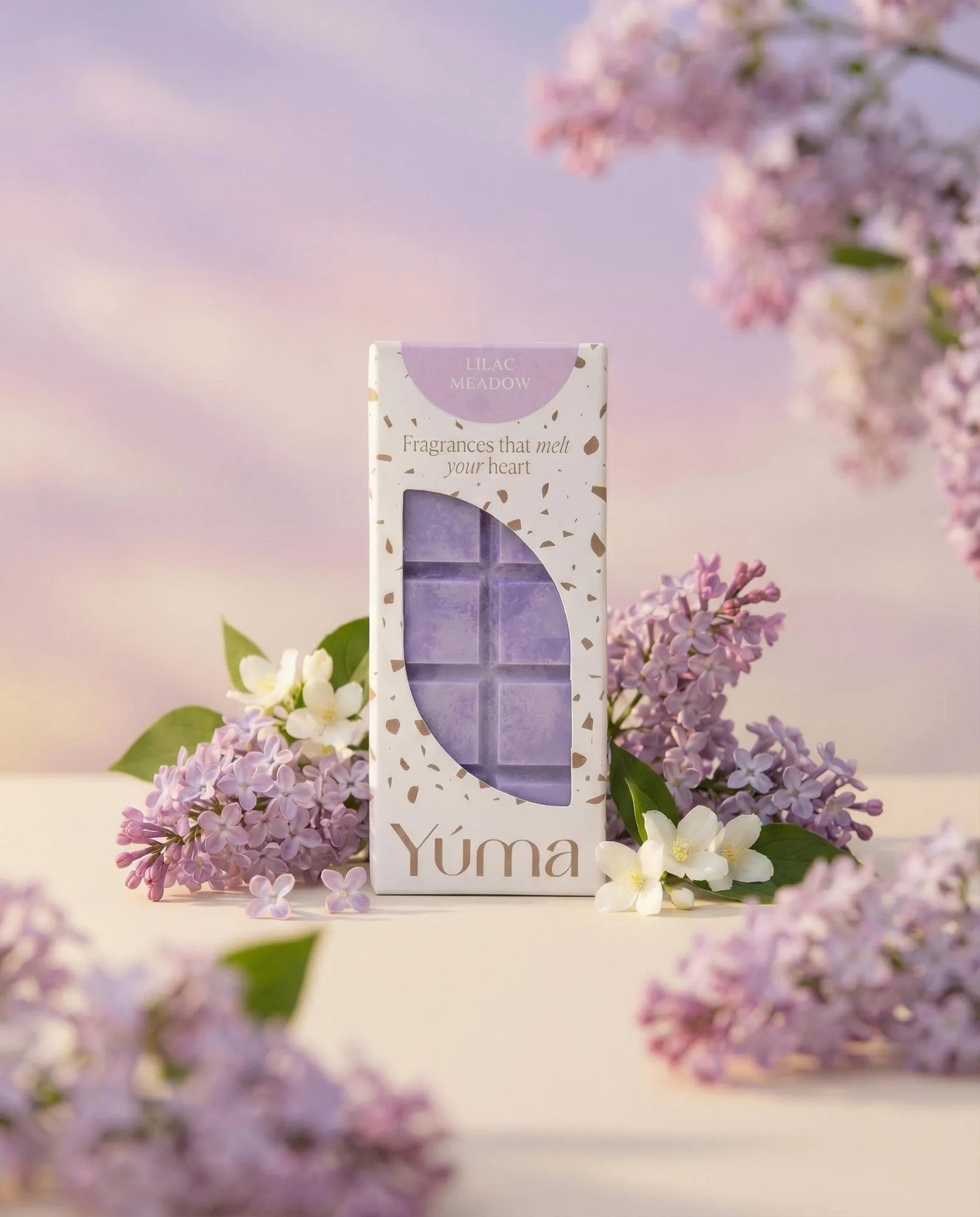 Μωβ wax melts Lilac Meadow της YUMA σε λευκή συσκευασία, περιτριγυρισμένα από φρέσκα άνθη πασχαλιάς.