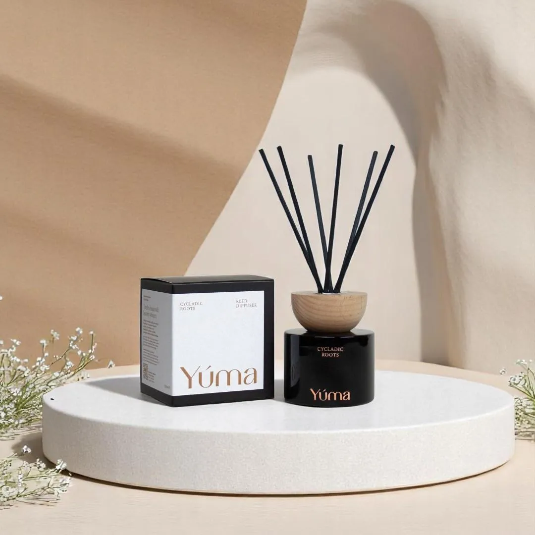 Το Cycladic Roots reed diffuser της YUMA με μαύρα στικς δίπλα στη συσκευασία του πάνω σε βάθρο.