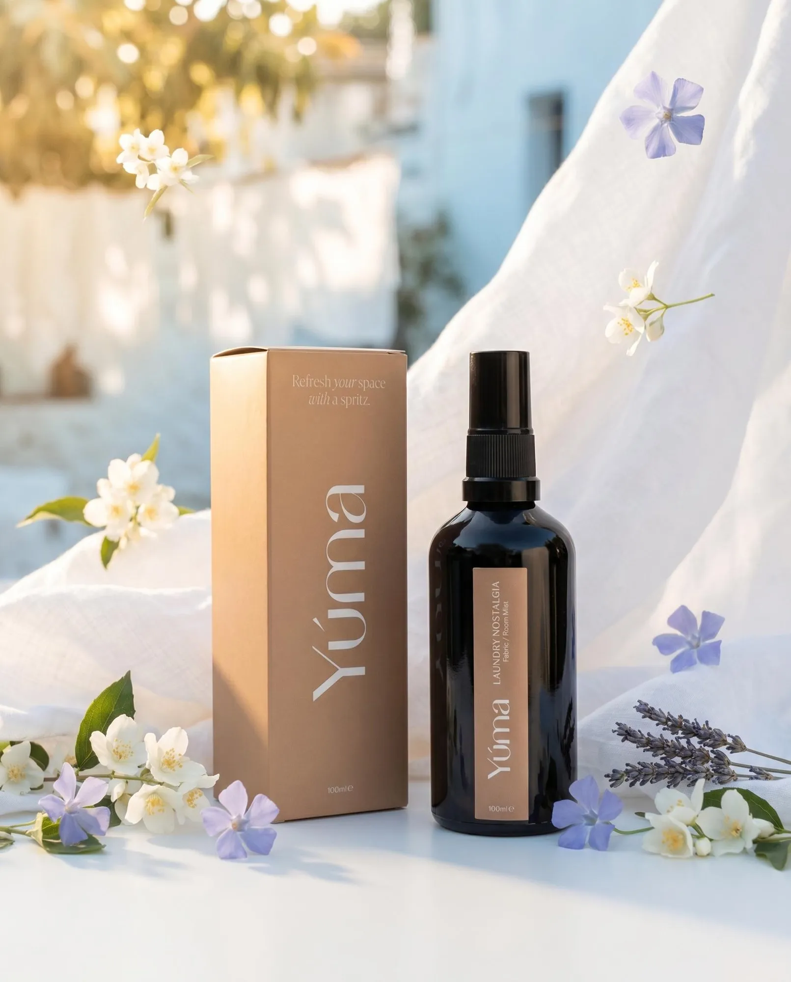 Το Laundry Nostalgia fabric mist της YUMA δίπλα στη συσκευασία του, περιτριγυρισμένο από λευκά και μωβ άνθη.