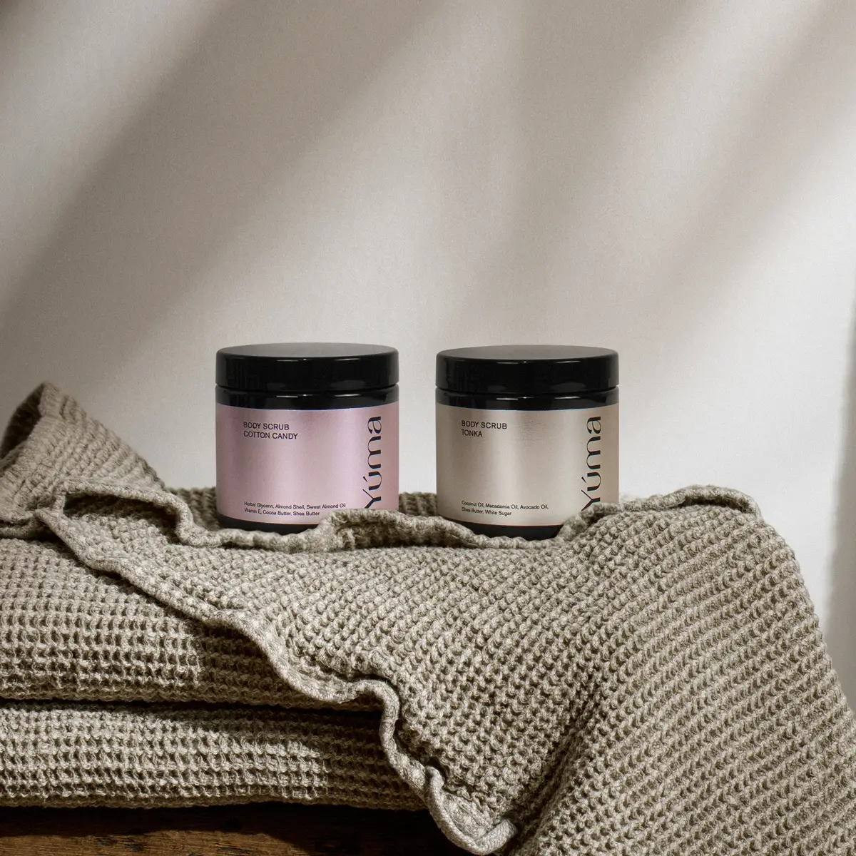 Δύο βάζα body scrub της YUMA, Cotton Candy και Tonka, πάνω σε πλεκτή πετσέτα Δύο βάζα body scrub της YUMA, Cotton Candy και Tonka, πάνω σε πλεκτή πετσέτα