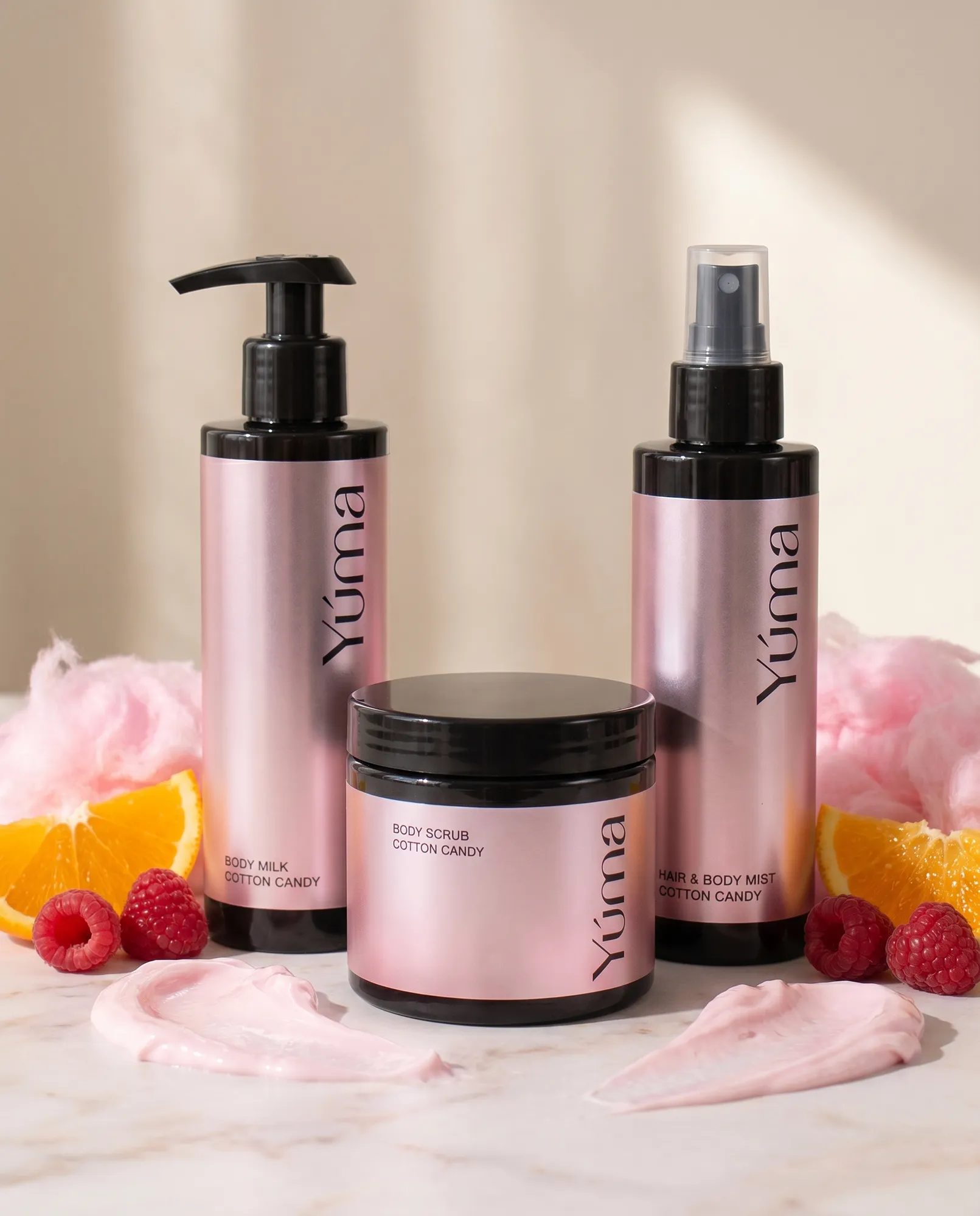 Cotton Candy Body Care Set Τρία ροζ μεταλλικά μπουκάλια Cotton Candy της YUMA ανάμεσα σε σμέουρα, πορτοκάλια και ροζ μαλλί της γριάς.