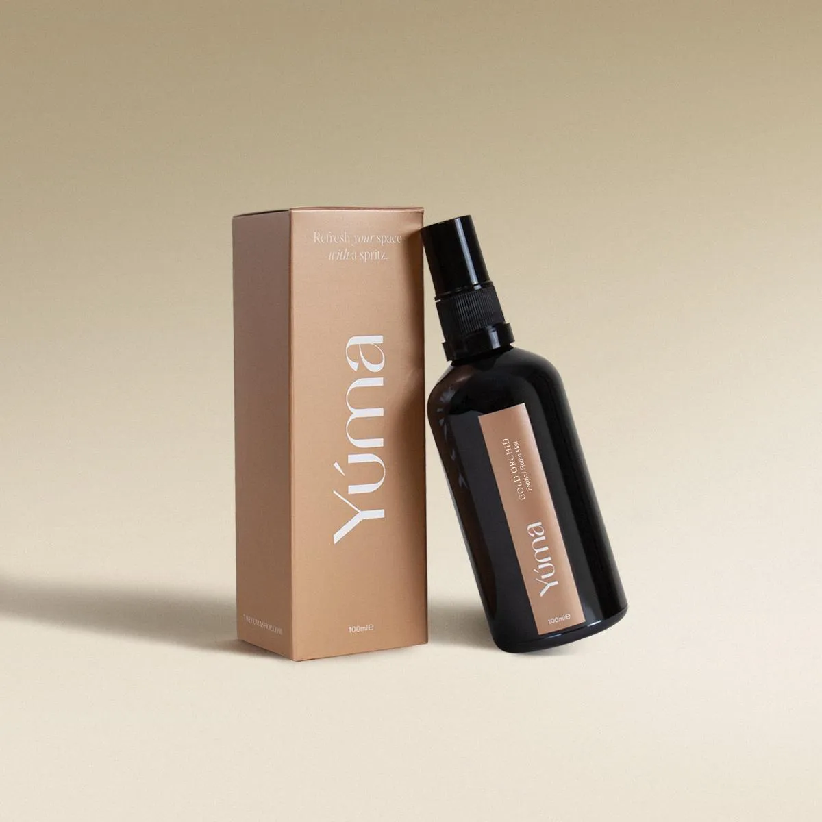 Gold Orchid Fabric/Room Mist Σκούρο γυάλινο μπουκάλι spray και χάρτινο κουτί της YUMA με το άρωμα Gold Orchid σε μπεζ φόντο.