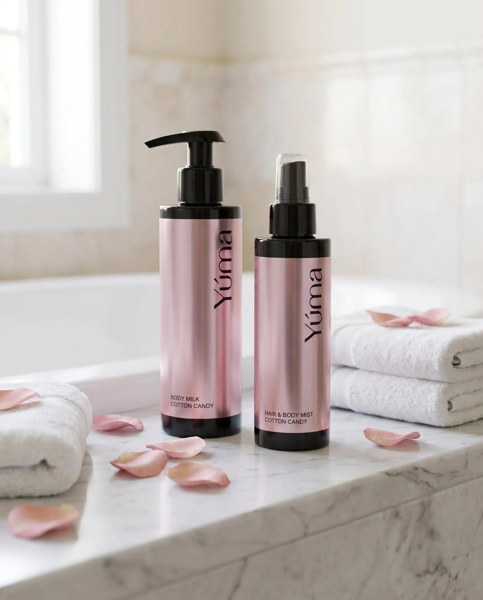 Body Milk & Δώρο Mist Cotton Candy Δύο ροζ μεταλλικά μπουκάλια Cotton Candy της YUMA δίπλα σε λευκές πετσέτες και ροδοπέταλα.