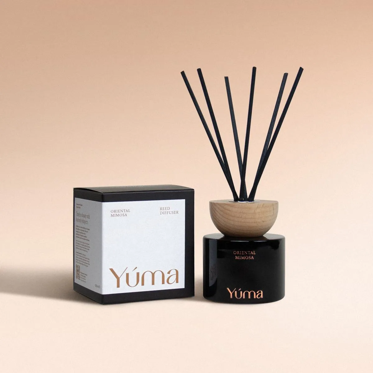 Oriental Mimosa Reed Diffuser Reed diffuser Oriental Mimosa της YUMA σε μαύρο γυάλινο δοχείο με ξύλινο πώμα δίπλα στη συσκευασία του.