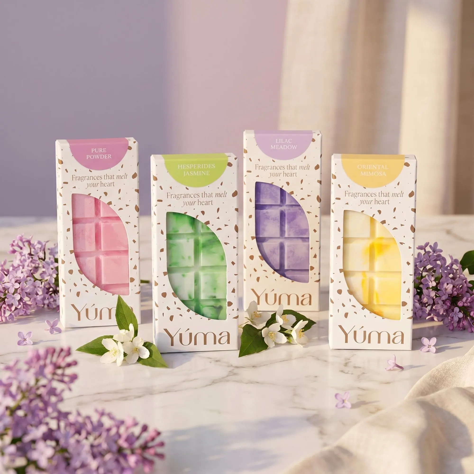 Signature Collection - Wax Melt Set Τέσσερα wax melts της YUMA σε χάρτινες συσκευασίες πάνω σε μάρμαρο με άνθη πασχαλιάς και γιασεμιού.