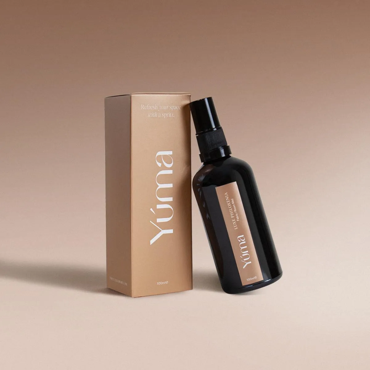Luxe Philoxenia Fabric/Room Mist Σκούρο γυάλινο μπουκάλι spray Luxe Philoxenia δίπλα στη χάρτινη μπεζ συσκευασία της YUMA σε ουδέτερο φόντο.