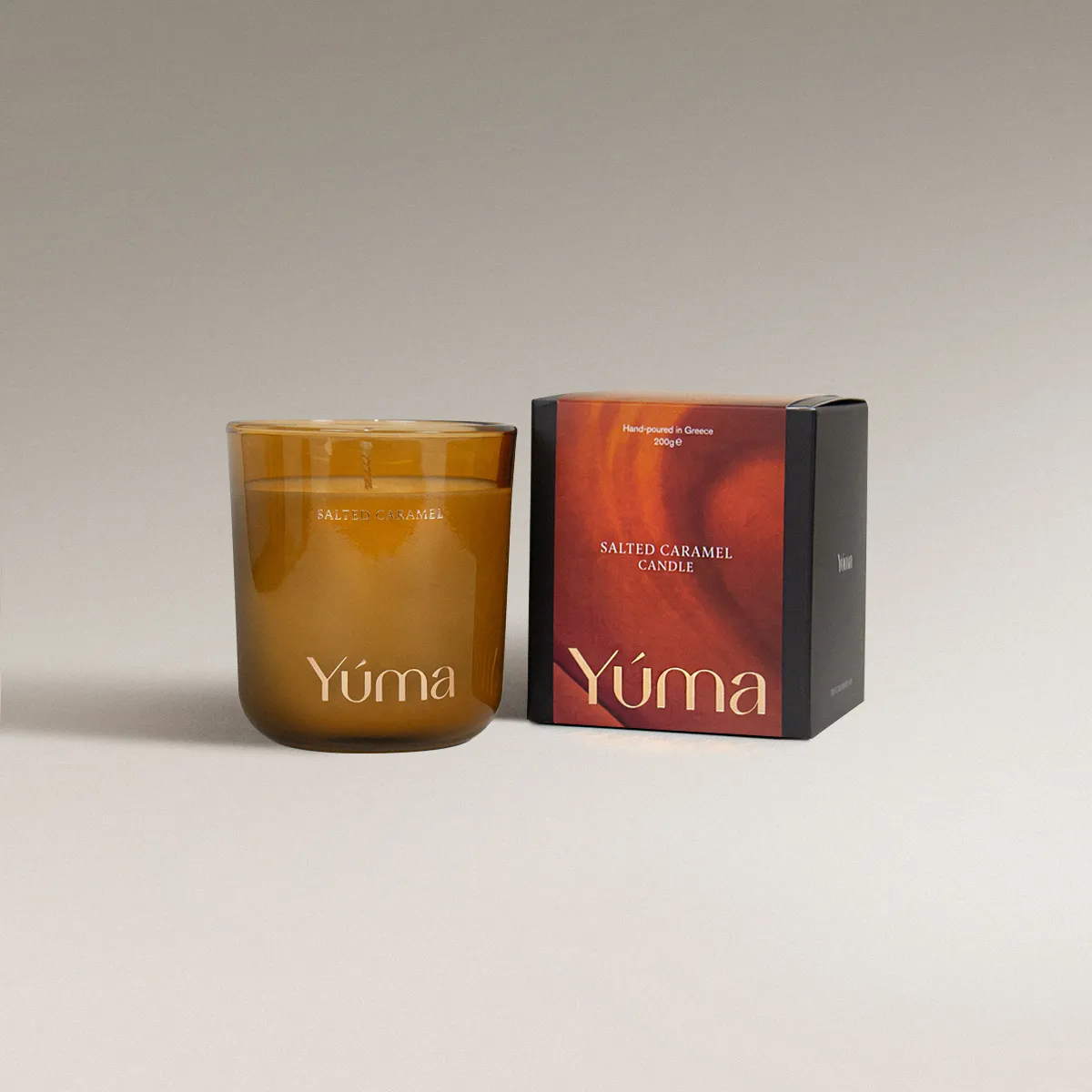 Salted Caramel Candle Κερί Salted Caramel της YUMA σε κεχριμπαρένιο γυάλινο δοχείο δίπλα στη χάρτινη συσκευασία του.