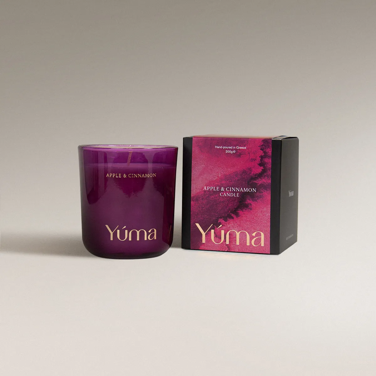 Apple Cinnamon Candle Κερί Apple Cinnamon της YUMA σε καπνιστό ροζ γυάλινο δοχείο δίπλα στη χάρτινη συσκευασία του.